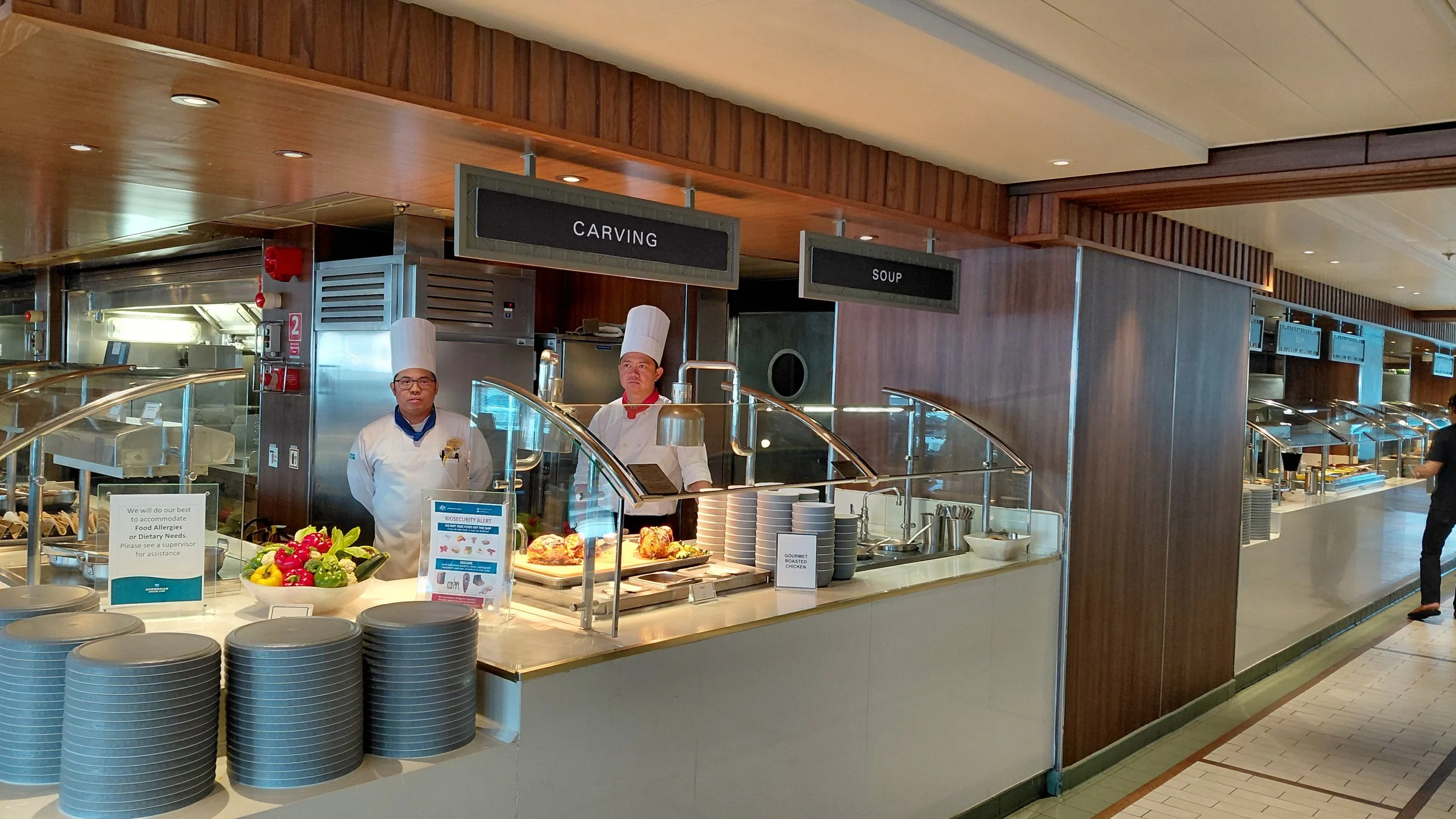 A buffet station in a restaurant or hotel with chefs standing behind the counter. There are two signs above the station, one reads 'Carving' and the other 'Soup.' Plates are stacked on the counter, and a bowl of colorful vegetables is visible in the 