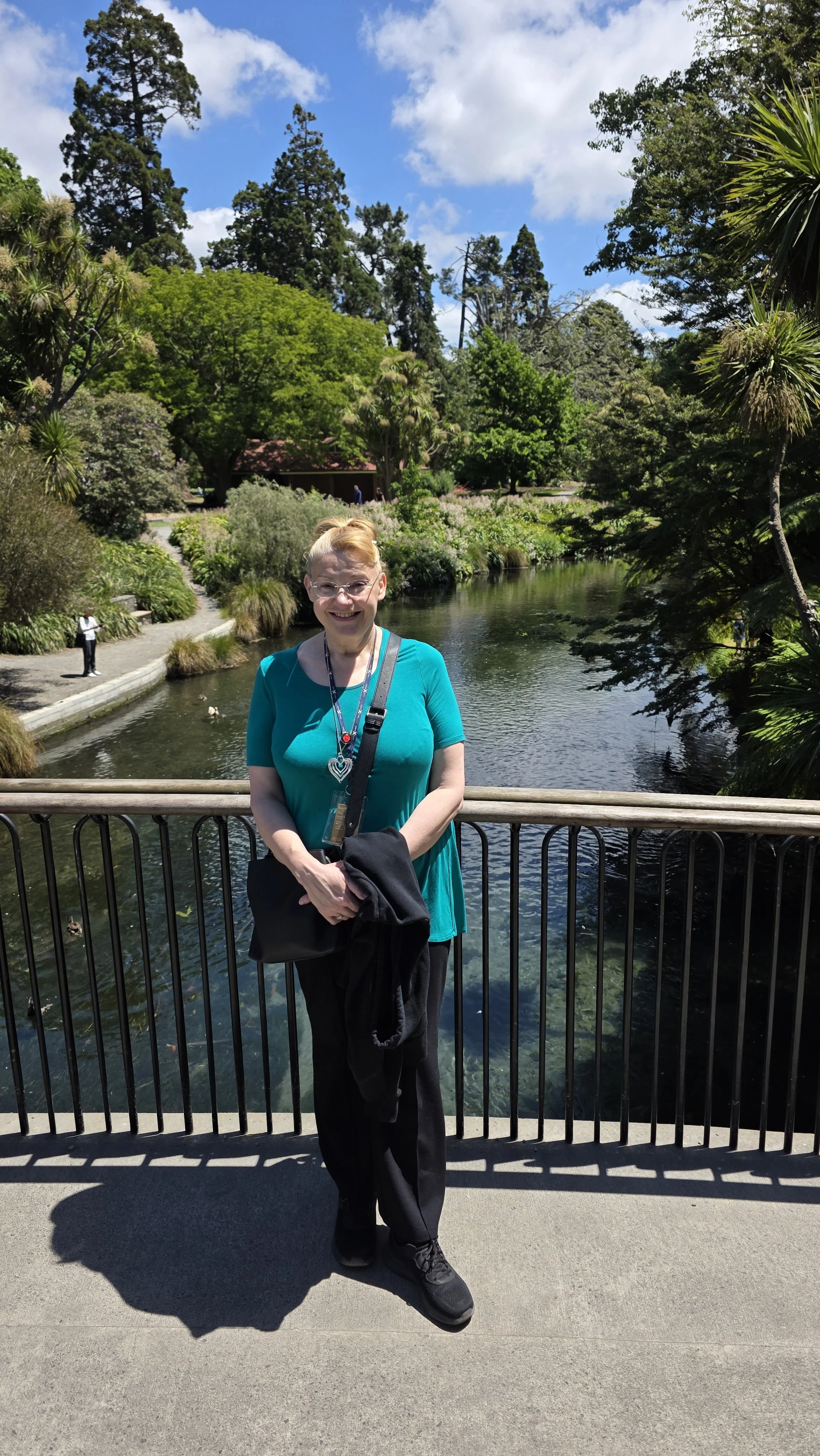 A woman with glasses and a cheerful smile, wearing a teal top and black pants, standing on a bridge in a lush park by a pond, under a partly cloudy sky.