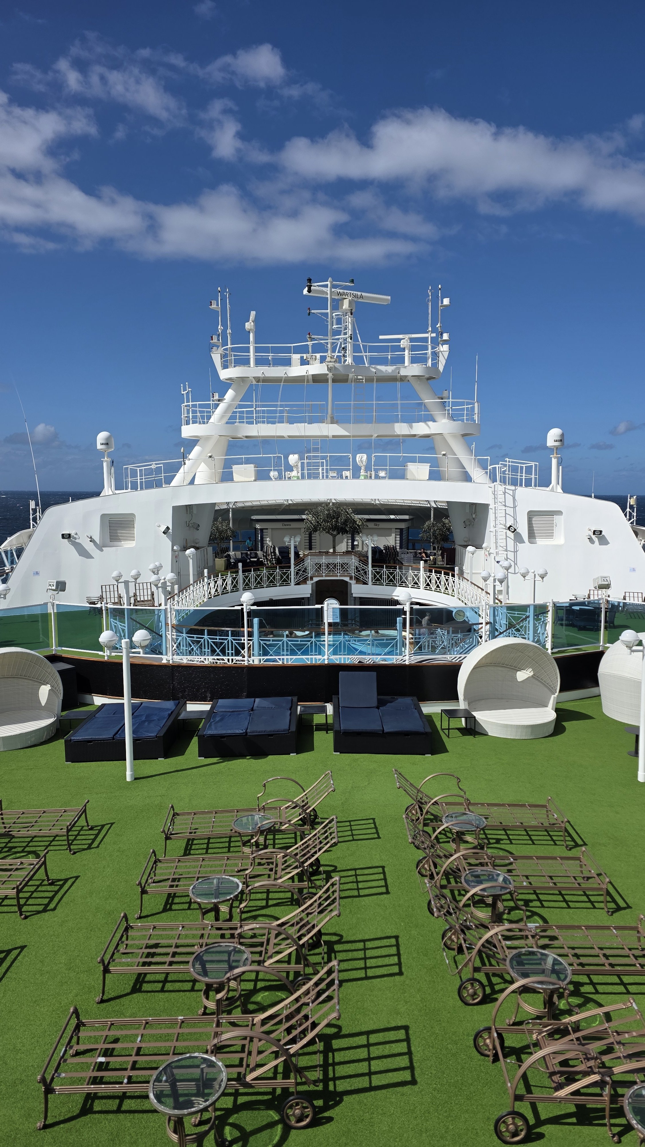 View of a cruise ship's outdoor deck with empty lounge chairs, round tables with chairs, green artificial grass, and a clear blue sky with a few clouds.