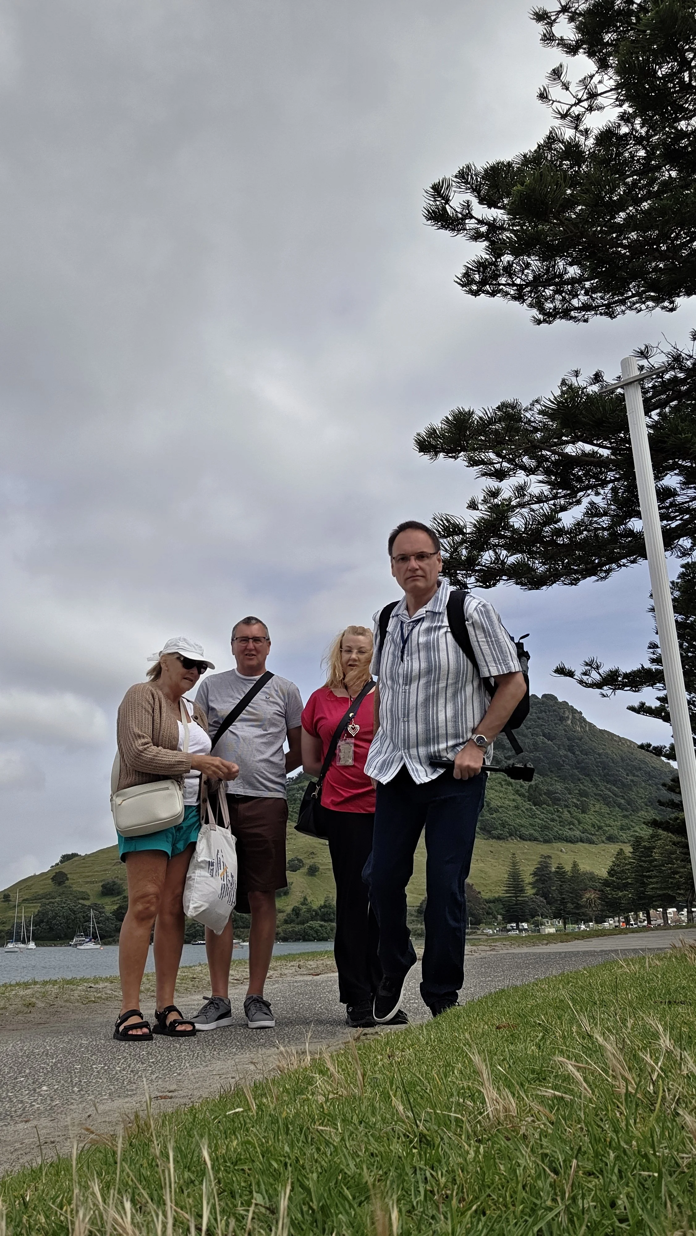 Four people standing outdoors near a body of water with boats, grassy area, trees, and hills in the background. They are dressed casually with backpacks and a camera.