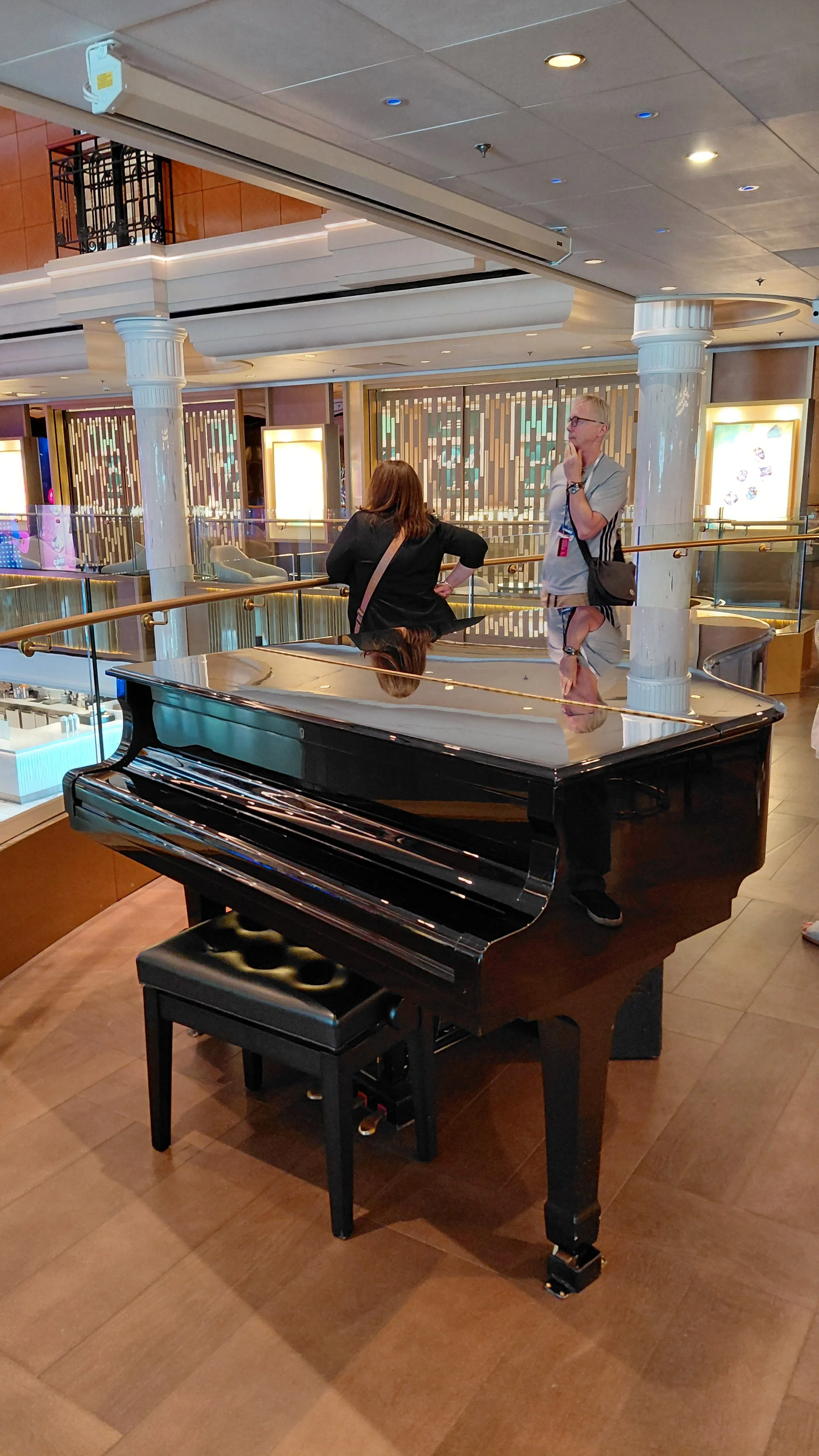A glossy black grand piano with a matching black bench in a shopping mall or hotel lobby. Two people, a woman and a man, are standing and having a conversation behind the piano near a railing. The background features modern decor with white columns, 