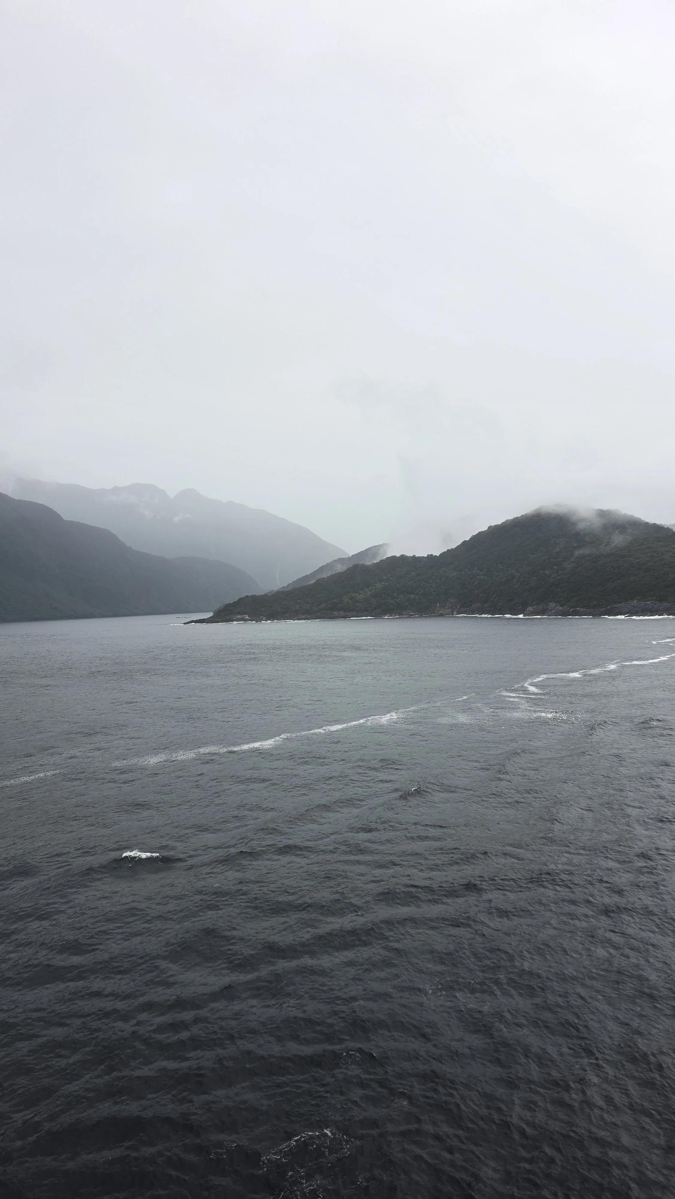 A scenic view of a cloudy sky over a body of water with forested hills and mountains in the background.