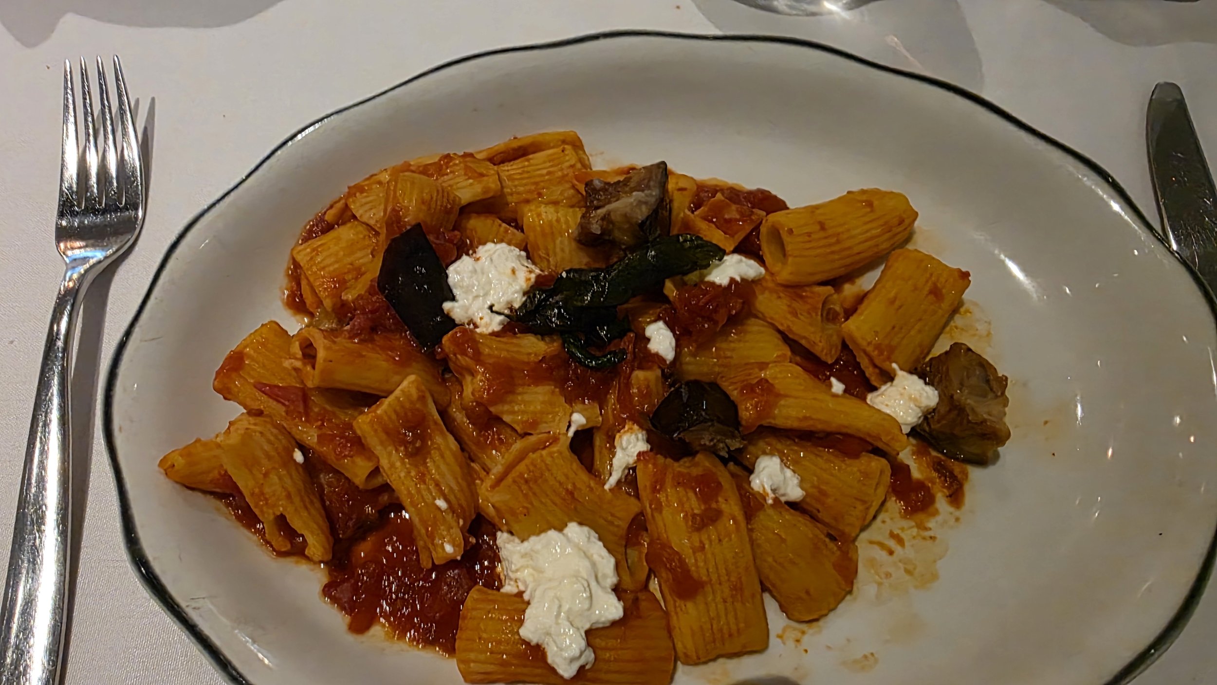 Plate of pasta with tomato sauce, topped with black olives, dollops of white cheese, served in a clear glass dish, with a fork on the left and a knife on the right.