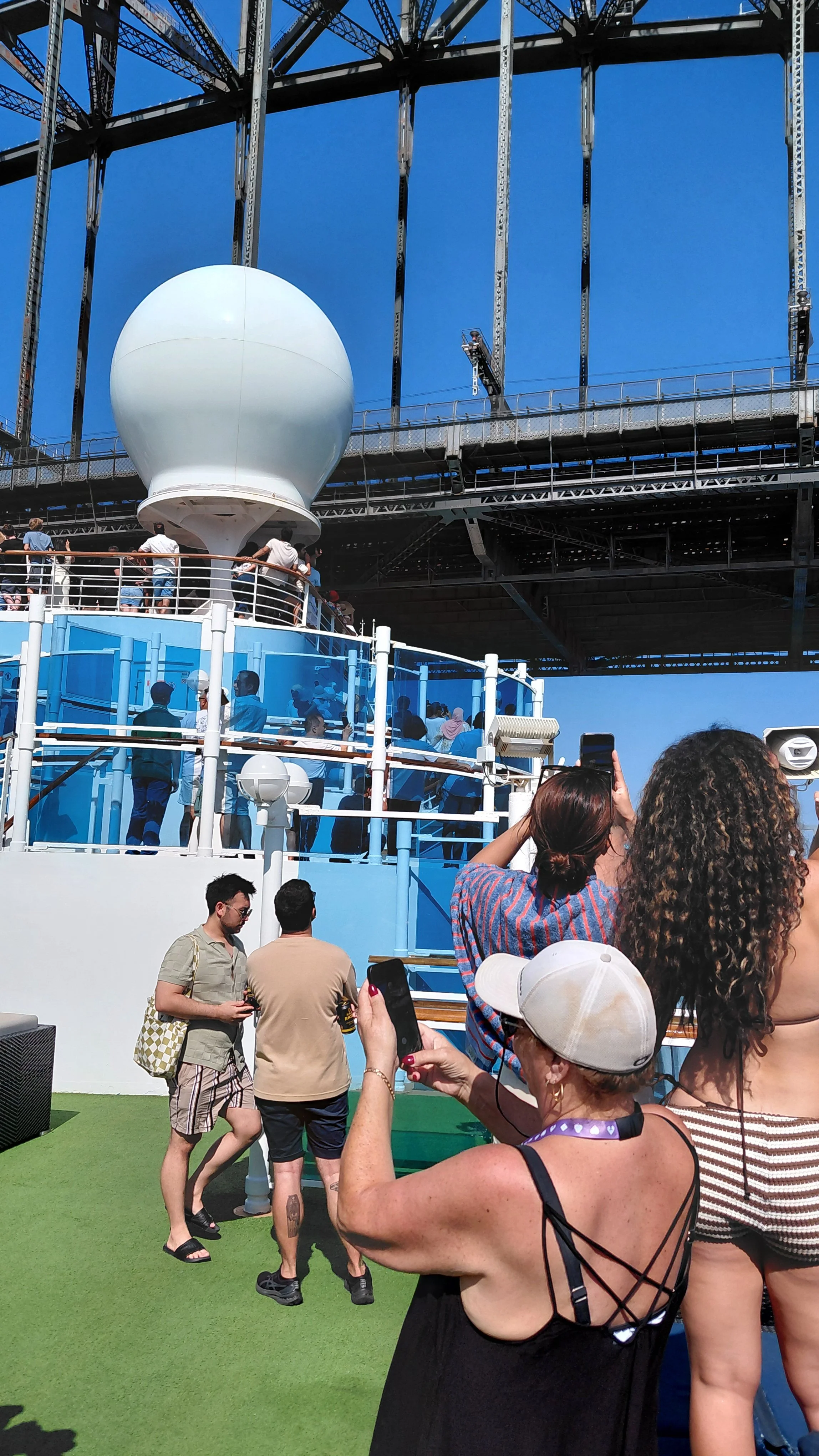 People standing on a cruise ship deck with a large white spherical radar and metal framework overhead against a clear blue sky.