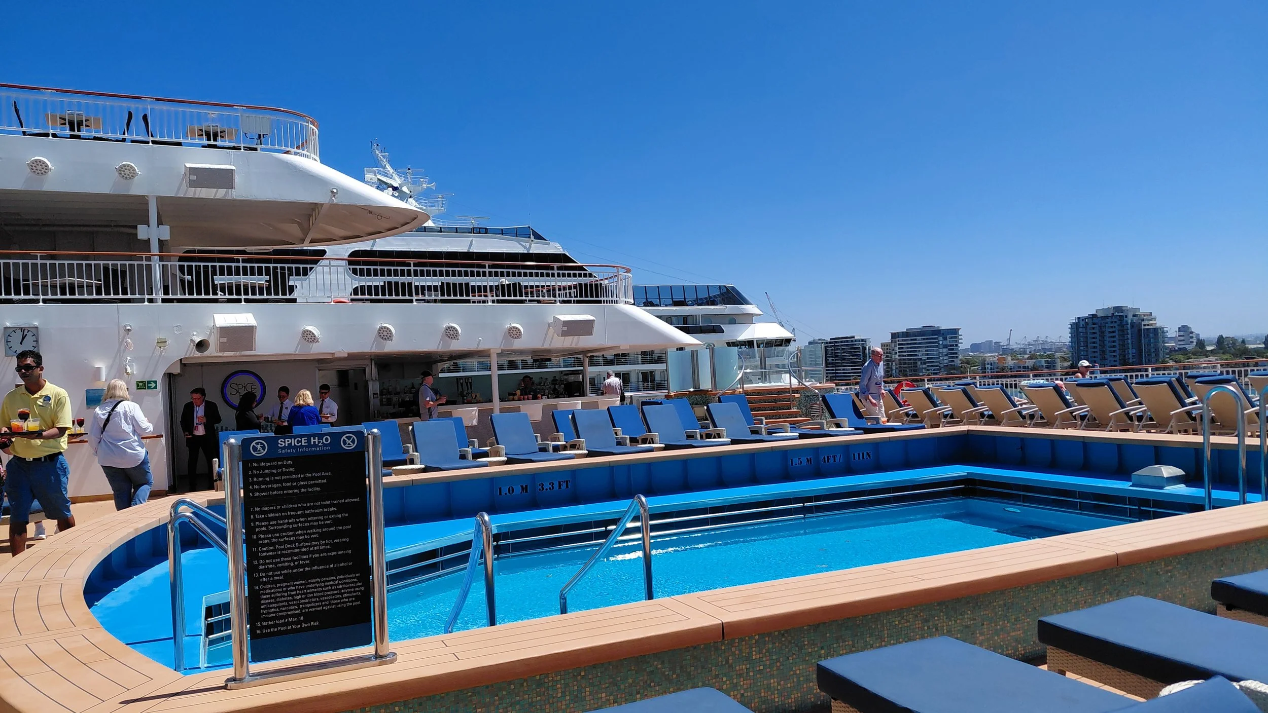 On the deck of a cruise ship, there is a small swimming pool surrounded by lounge chairs, with a city skyline in the background and a clear blue sky overhead.