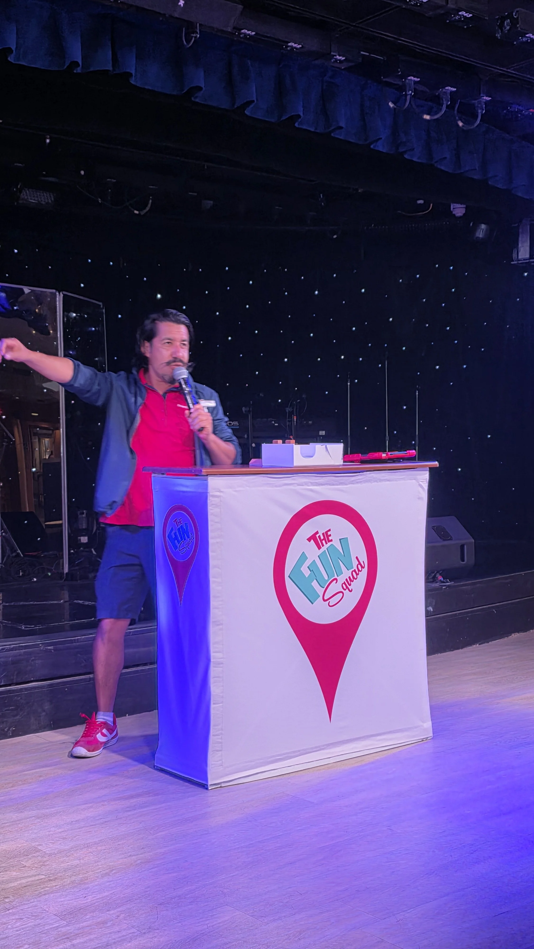 A man standing behind a podium with a microphone in a stage setting. The podium has a logo that reads 'The Fun Squad'. The man is wearing a red shirt, dark shorts, a blue jacket, and red sneakers.