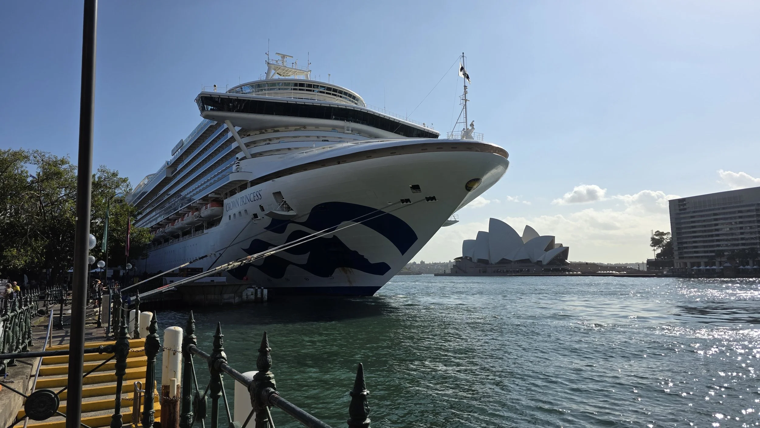 Large cruise ship docked at a harbor with the Sydney Opera House in the background and clear blue sky.