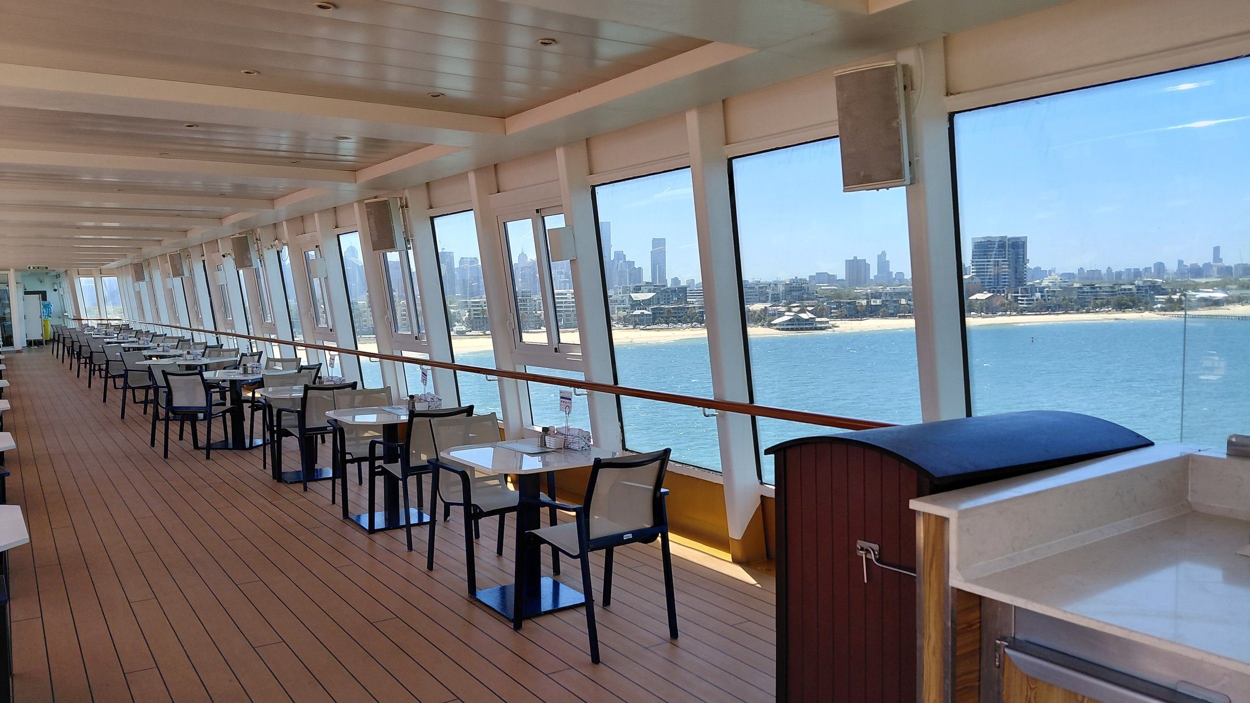 Interior of a water-view restaurant or lounge area with white tables and chairs lined up along large windows overlooking a body of water and a city skyline in the distance.