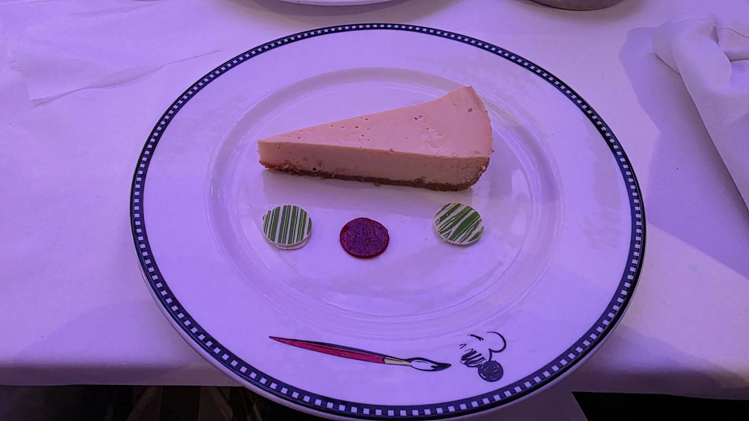 A slice of cheesecake on a plate decorated with green and white-striped candies and a red round candy, with a chef and spoon graphic around the rim of the plate.