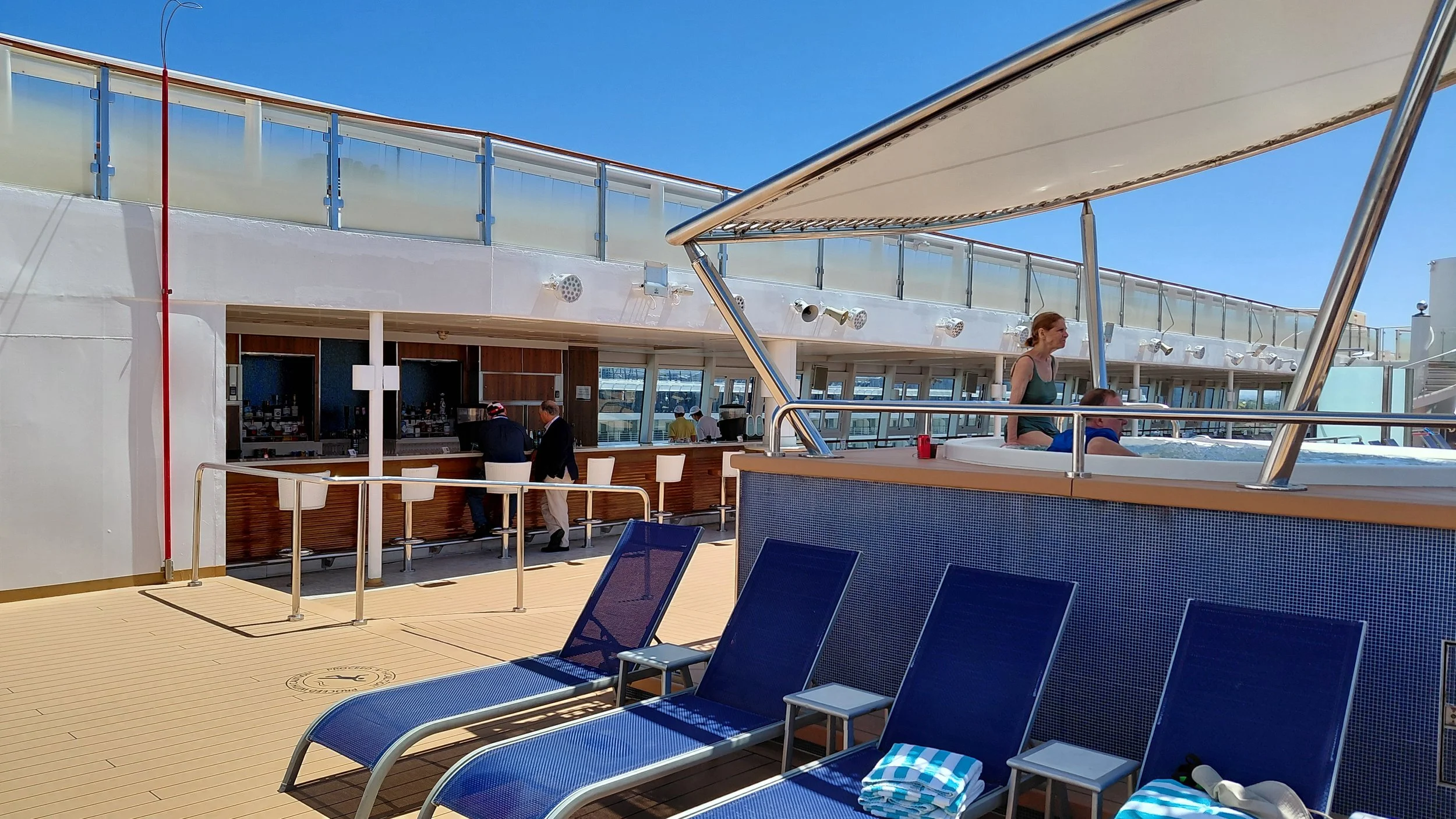 Empty blue lounge chairs with striped towels on a cruise ship deck, a hot tub with people, and a bar area in the background under a bright blue sky.