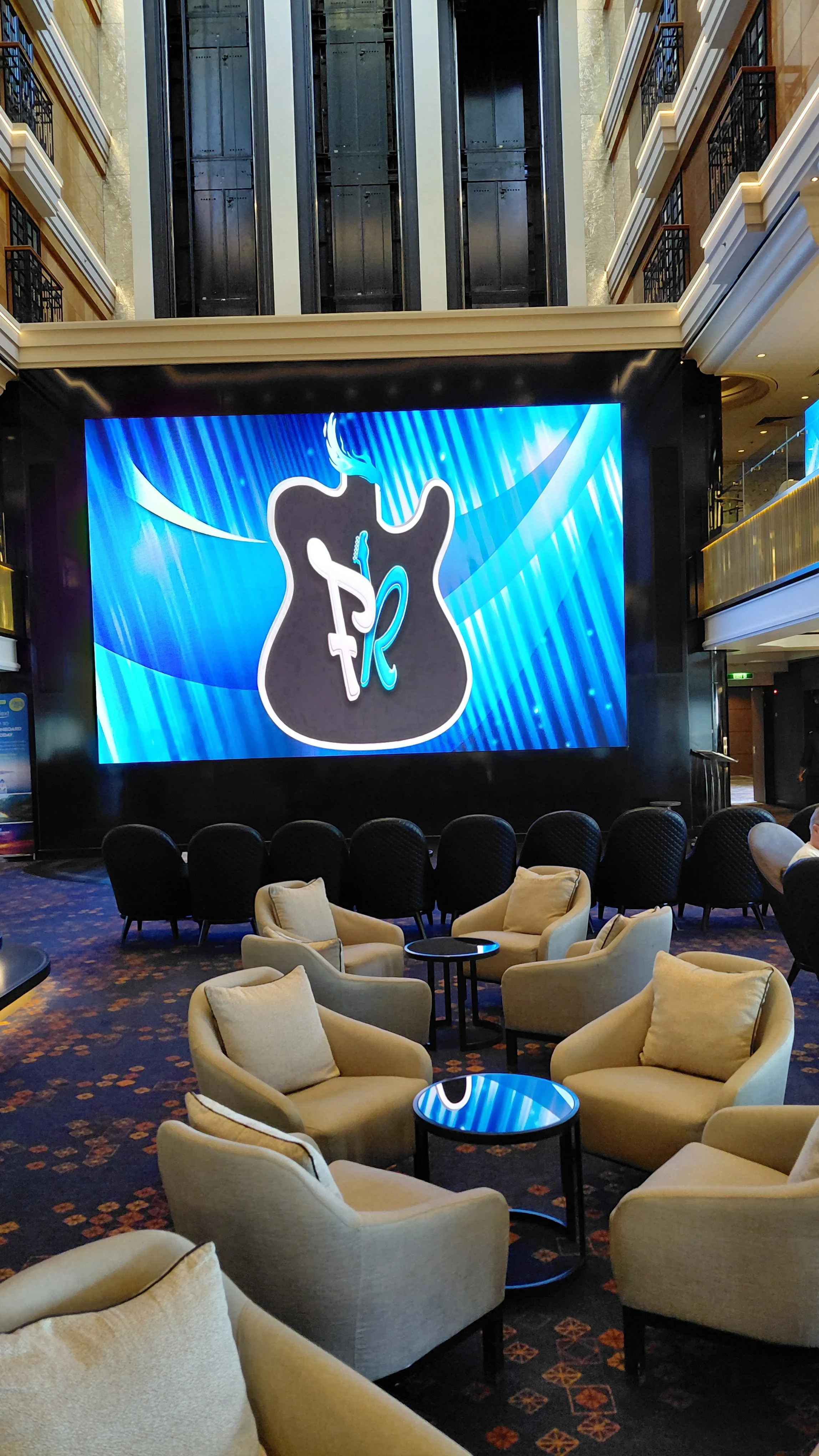 Interior of a modern cruise ship atrium with a large screen displaying a guitar icon and a cocktail glass and a smoking pipe; seating includes beige armchairs with pillows and black chairs, with a patterned carpet and multiple balconies with railings
