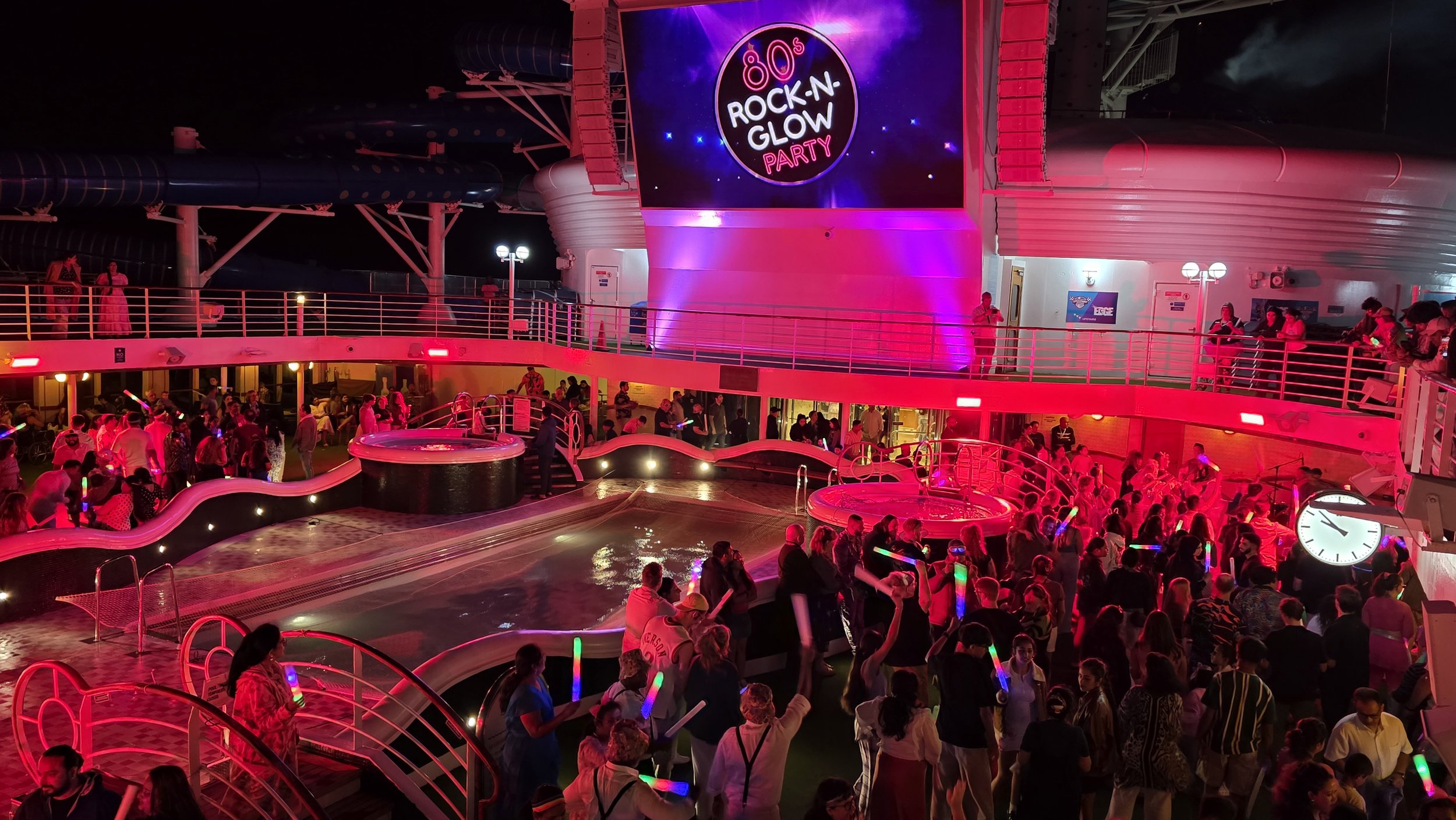 Crowd of people enjoying a glow party on a cruise ship at night with colorful lights, glow sticks, a swimming pool, and a large digital screen displaying a '80s Rock-N-Grow Party' advertisement.