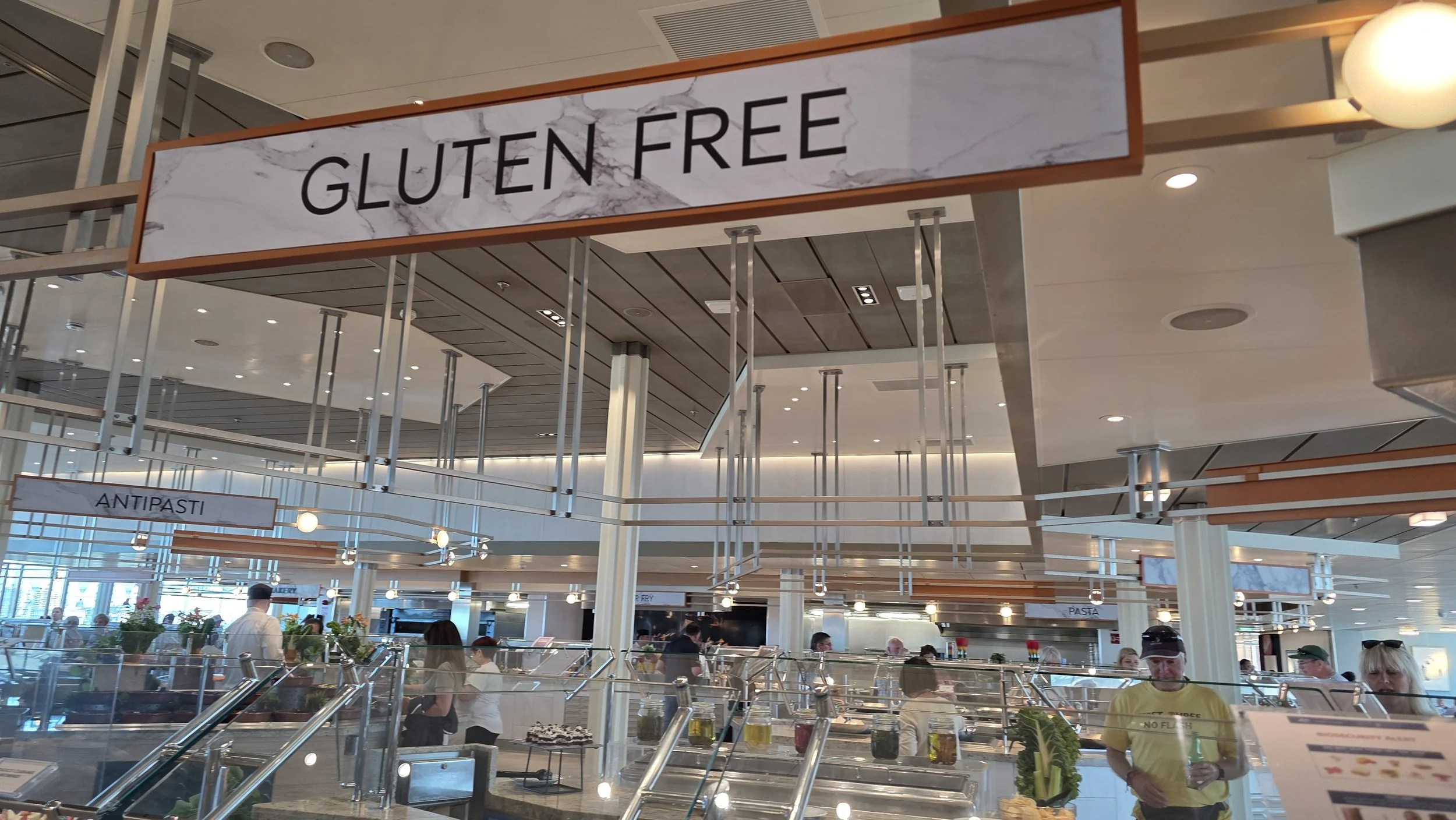 Restaurant buffet area with a sign that says "Gluten Free" and other signs for antipasti and pasta, with people serving themselves and food containers.
