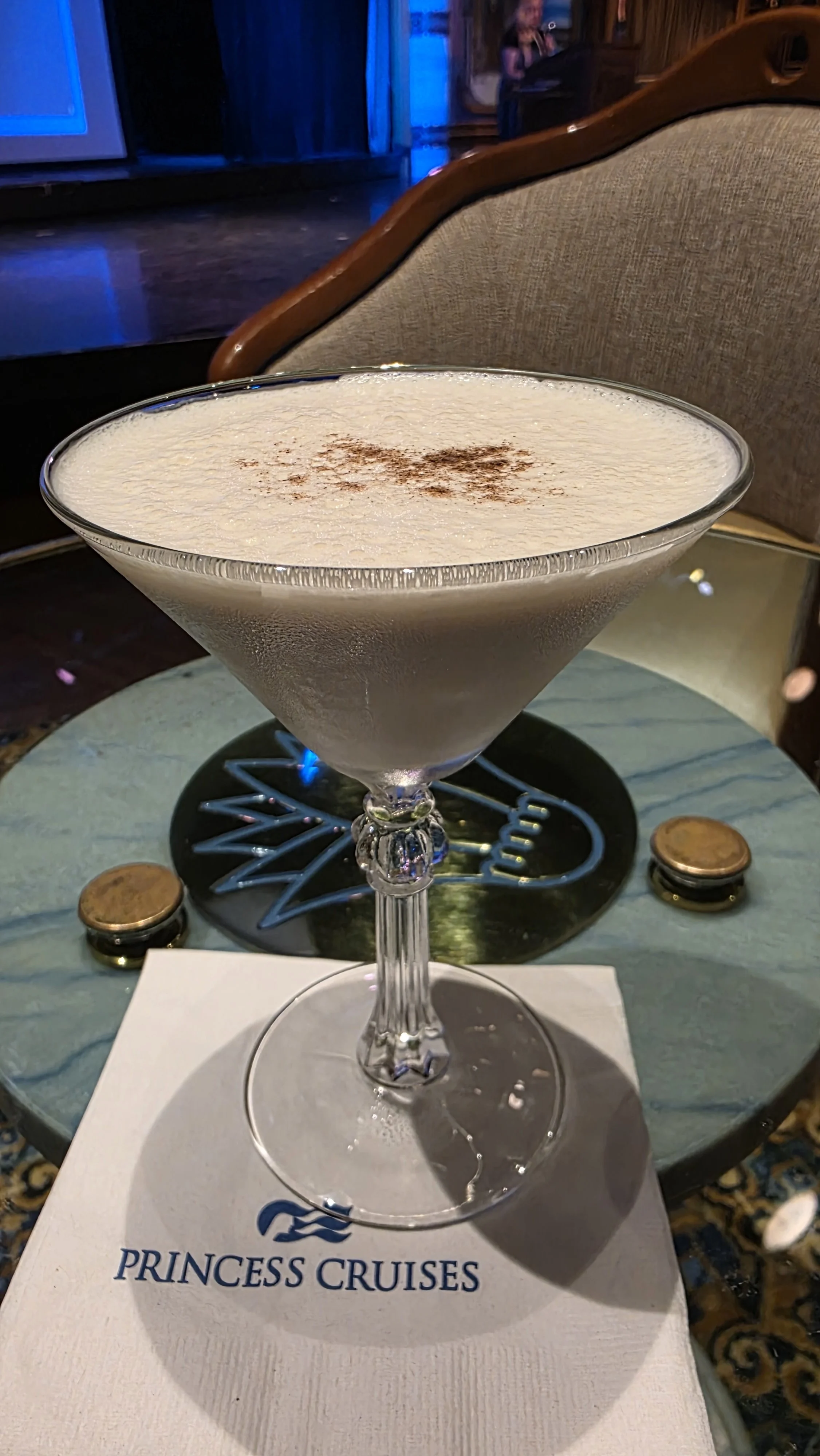 A cocktail drink in a martini glass with a frothy top, garnished with a sprinkle of cinnamon or cocoa powder, on a table with a Princess Cruises napkin.