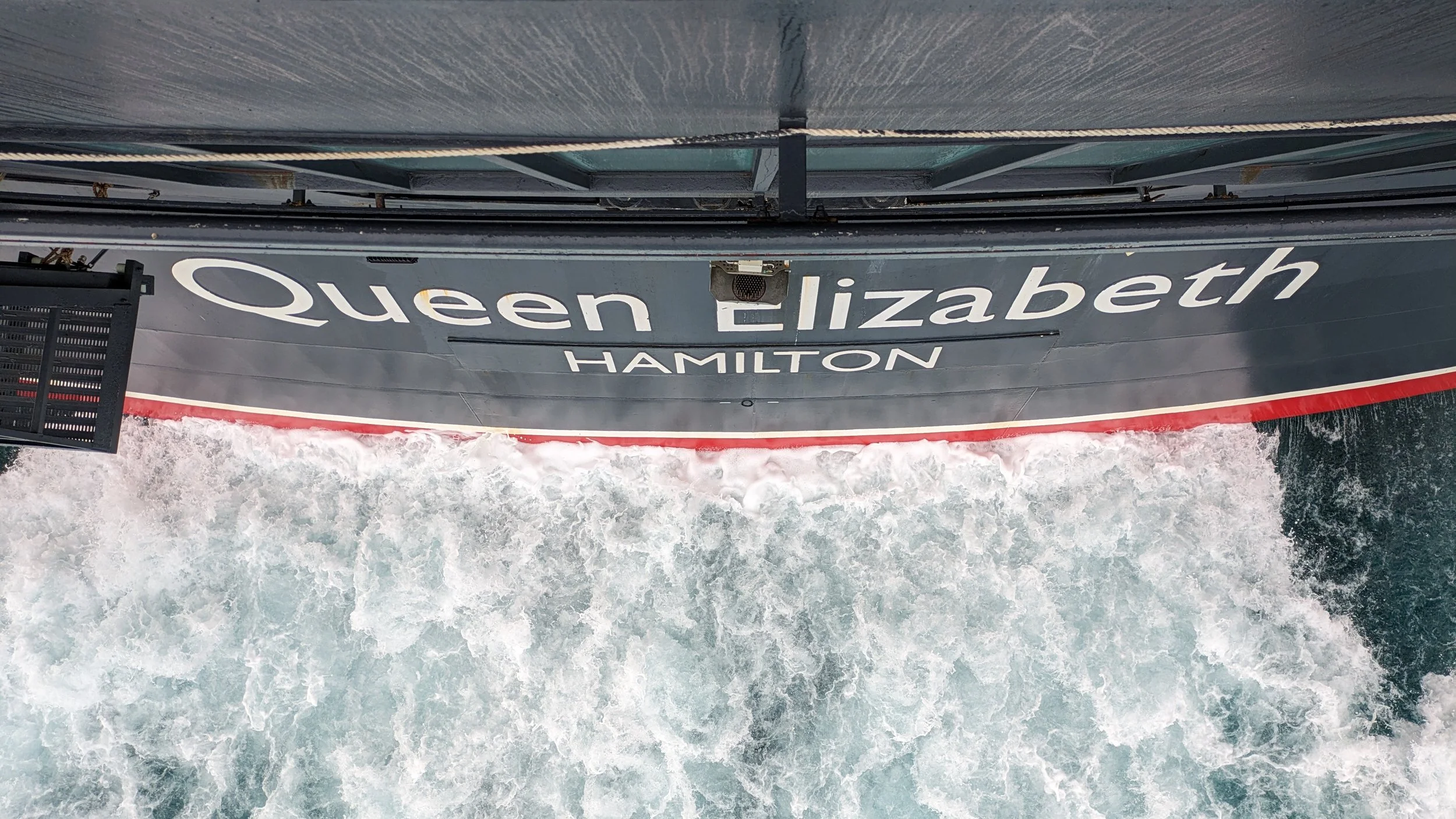 View from above of a boat labeled 'Queen Elizabeth Hamilton' moving through water, creating waves behind it.