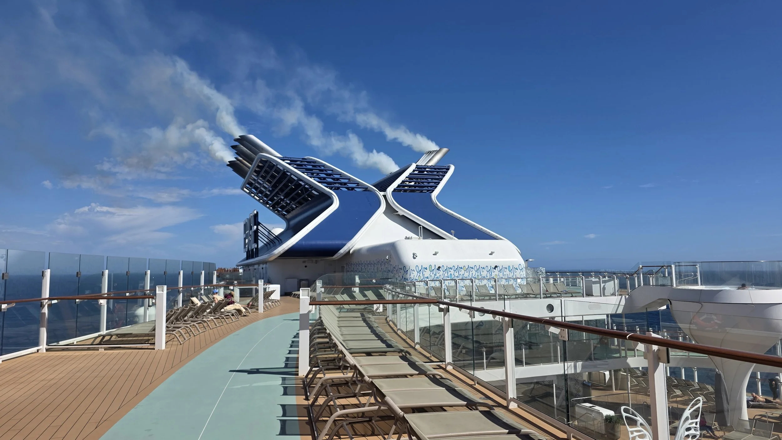 On the deck of a cruise ship, the ship's funnel with a modern design grows from the deck with glass railings, wooden benches, and lounge chairs, under a clear blue sky.
