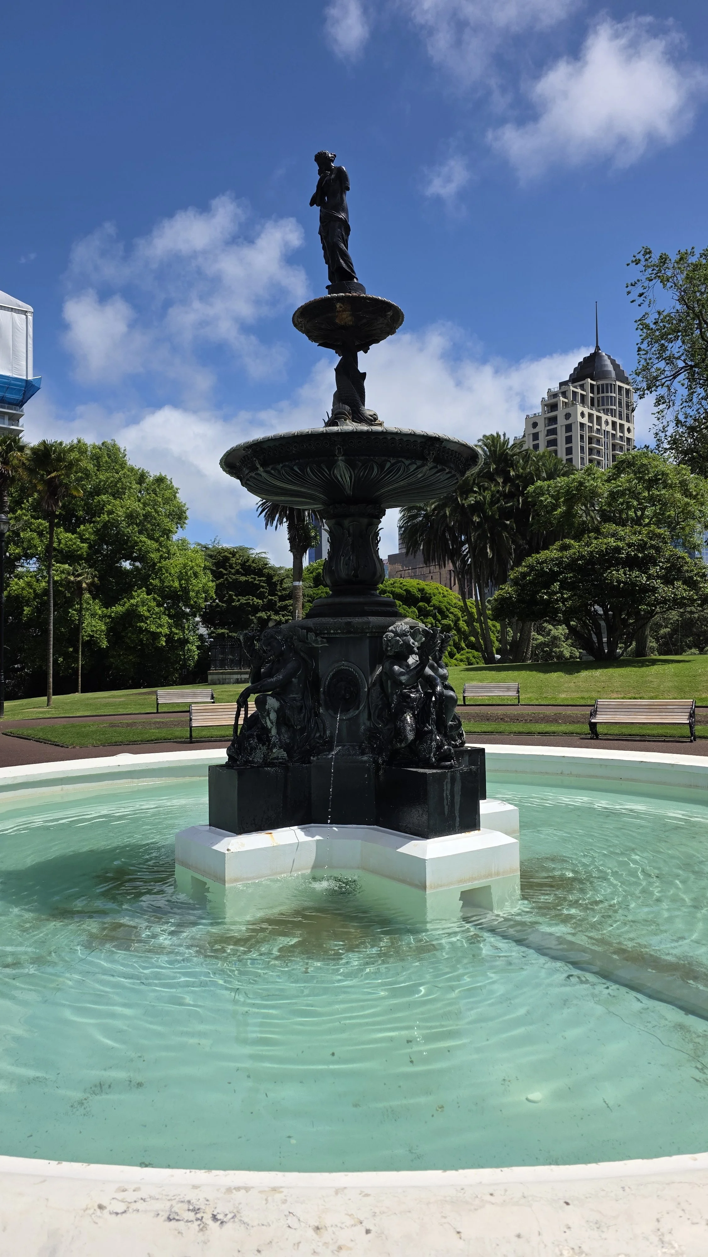 A decorative black fountain with sculptures, water flowing from the statues, in a park with green trees and benches, tall buildings in the background, under a blue sky with clouds.