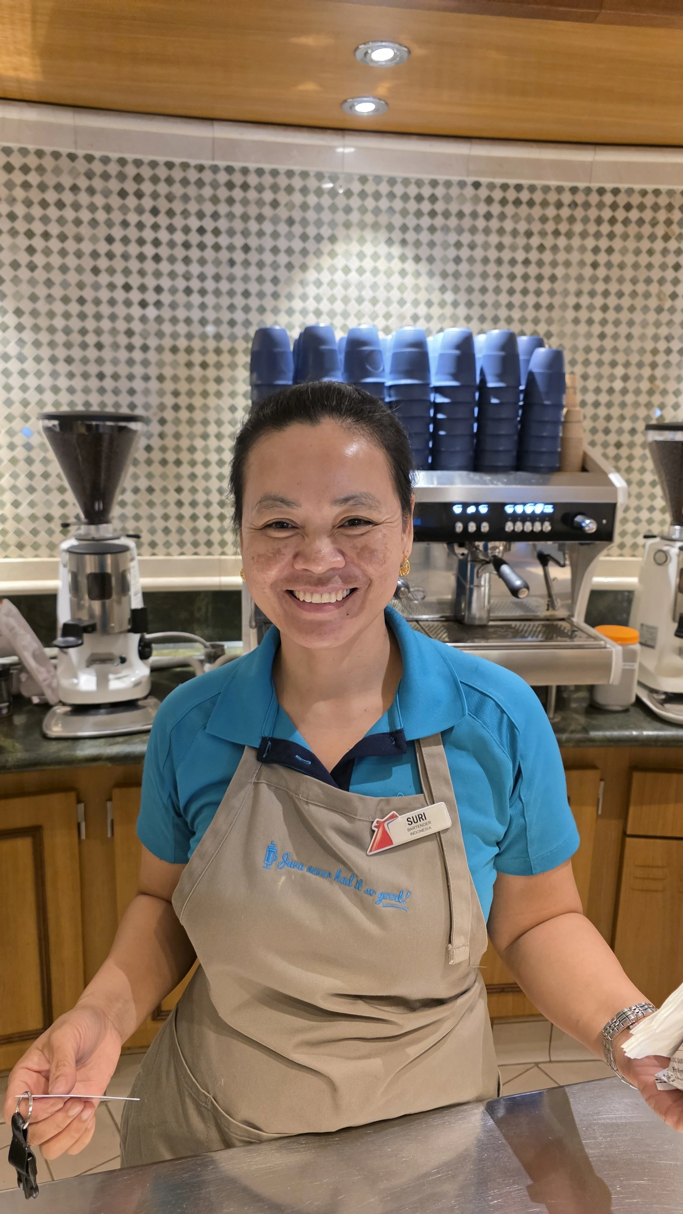 A smiling woman wearing a teal polo shirt and a beige apron standing behind a countertop at a coffee shop with coffee equipment and stacked blue cups in the background.