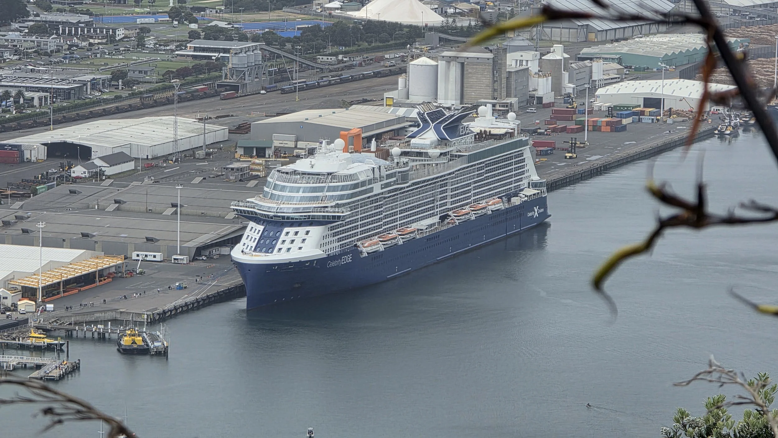 A large cruise ship named Celebrity EDGE docked at a port, with warehouses and industrial buildings nearby and a body of water in the foreground.