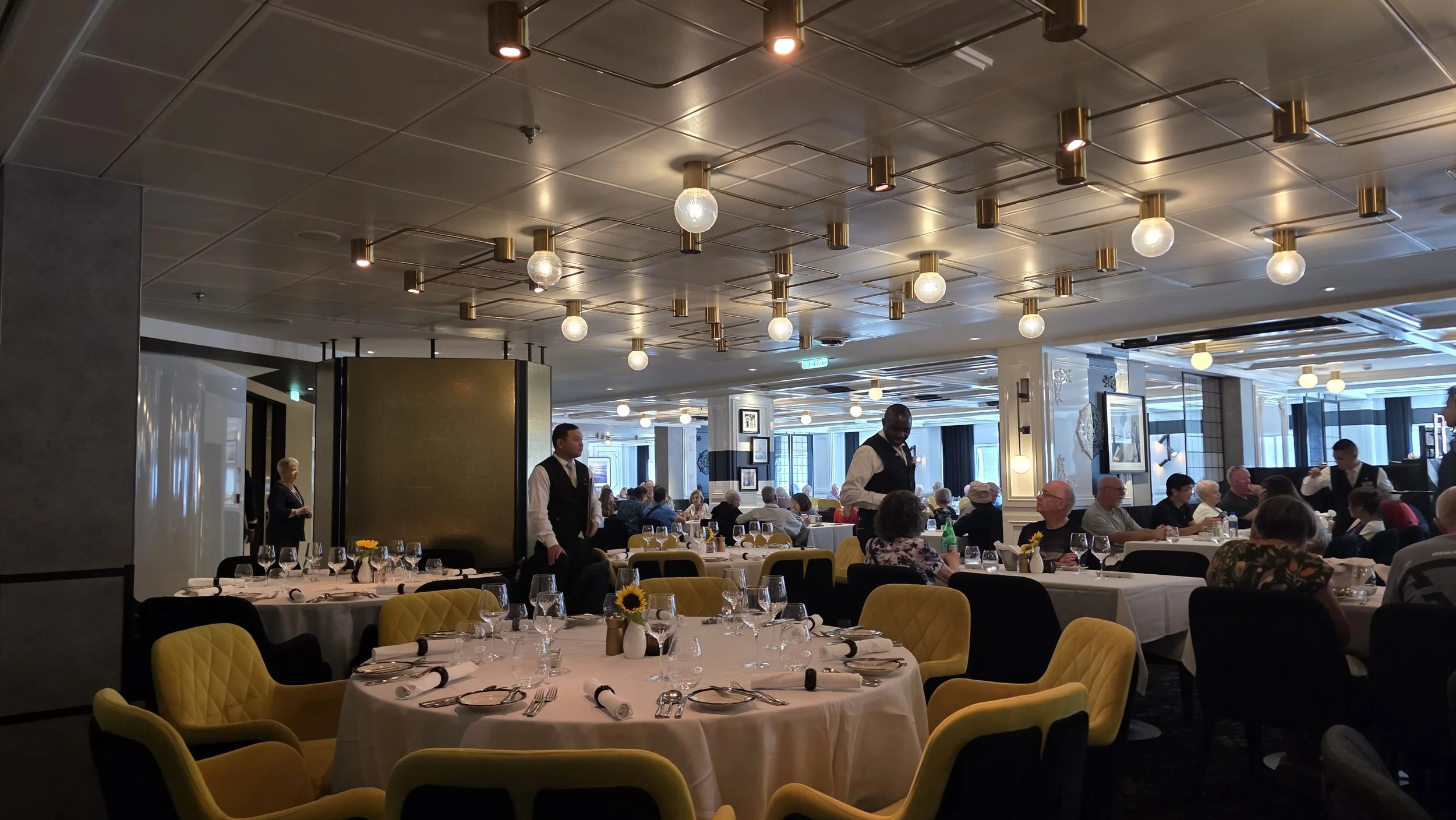 Elegant restaurant interior with round tables set with white tablecloths, glassware, plates, and napkins, surrounded by upholstered yellow and black chairs, and waitstaff attending to guests during daytime.