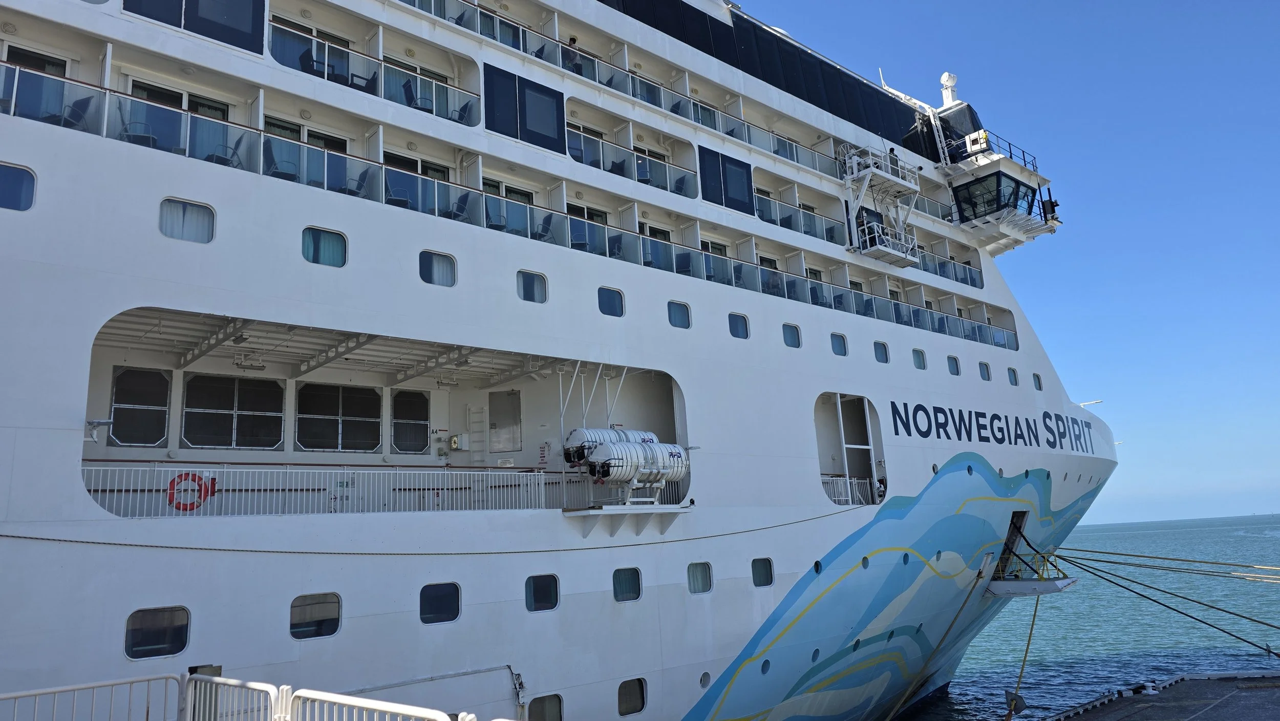 Large white cruise ship named Norwegian Spirit docked at the port, showing multiple decks, windows, balconies, and the ship's name on the side, with a blue sky and ocean in the background.