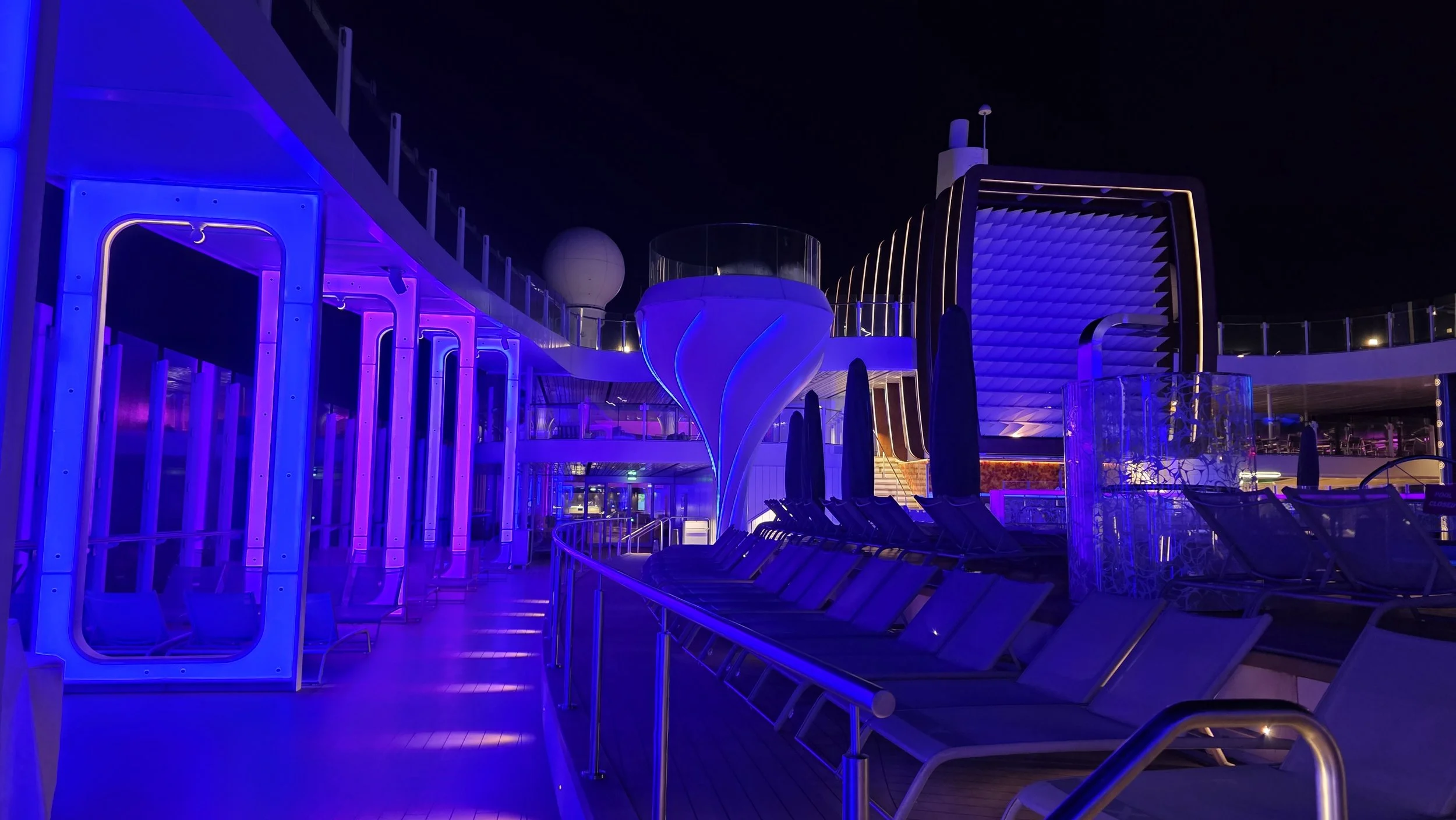 Empty cruise ship deck at night with blue and purple lighting, lounge chairs, and modern architectural structures.