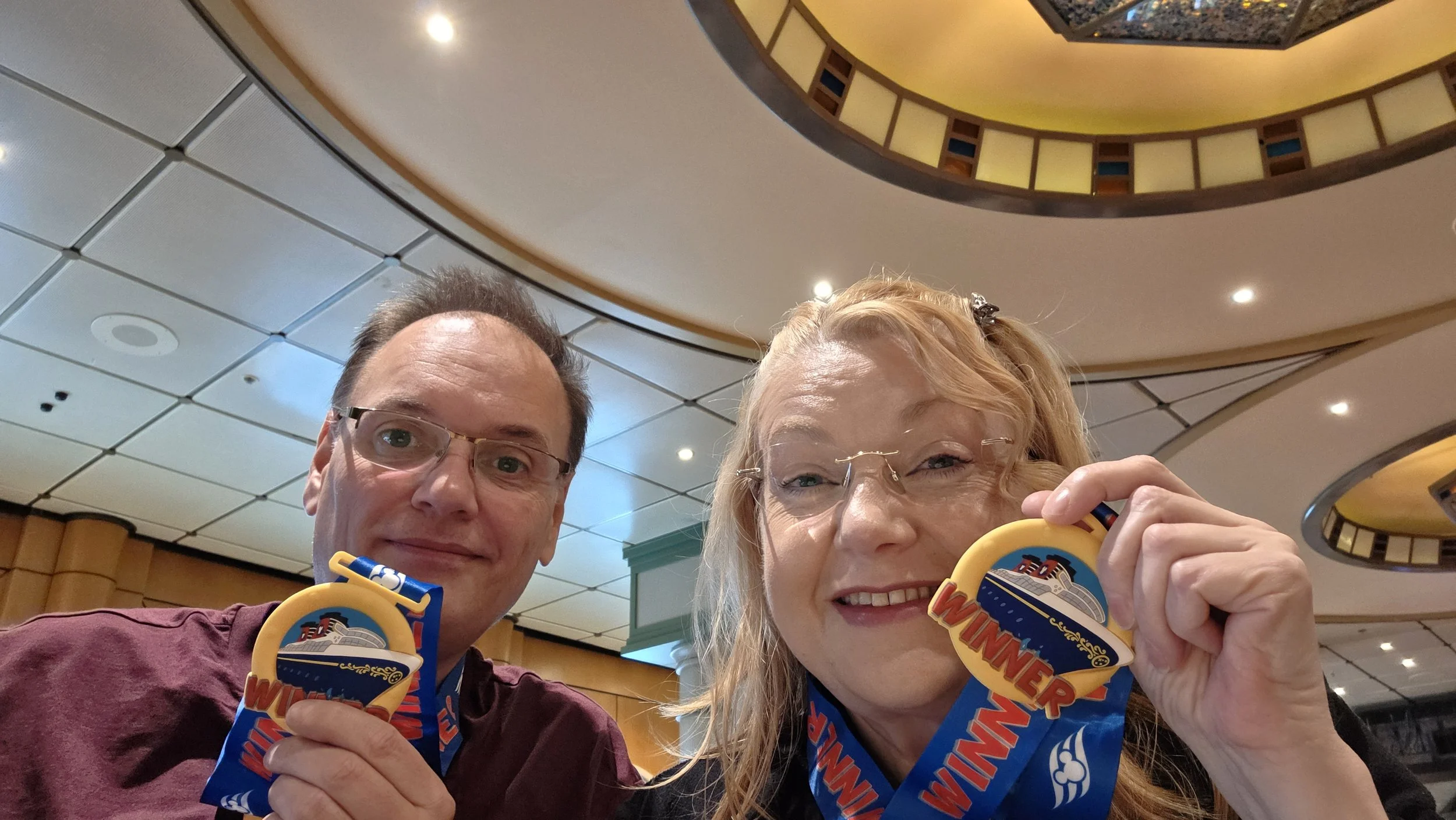 A man and woman hold up cruise ship medals, smiling for a selfie inside a decorated venue with circular ceiling design and warm lighting.