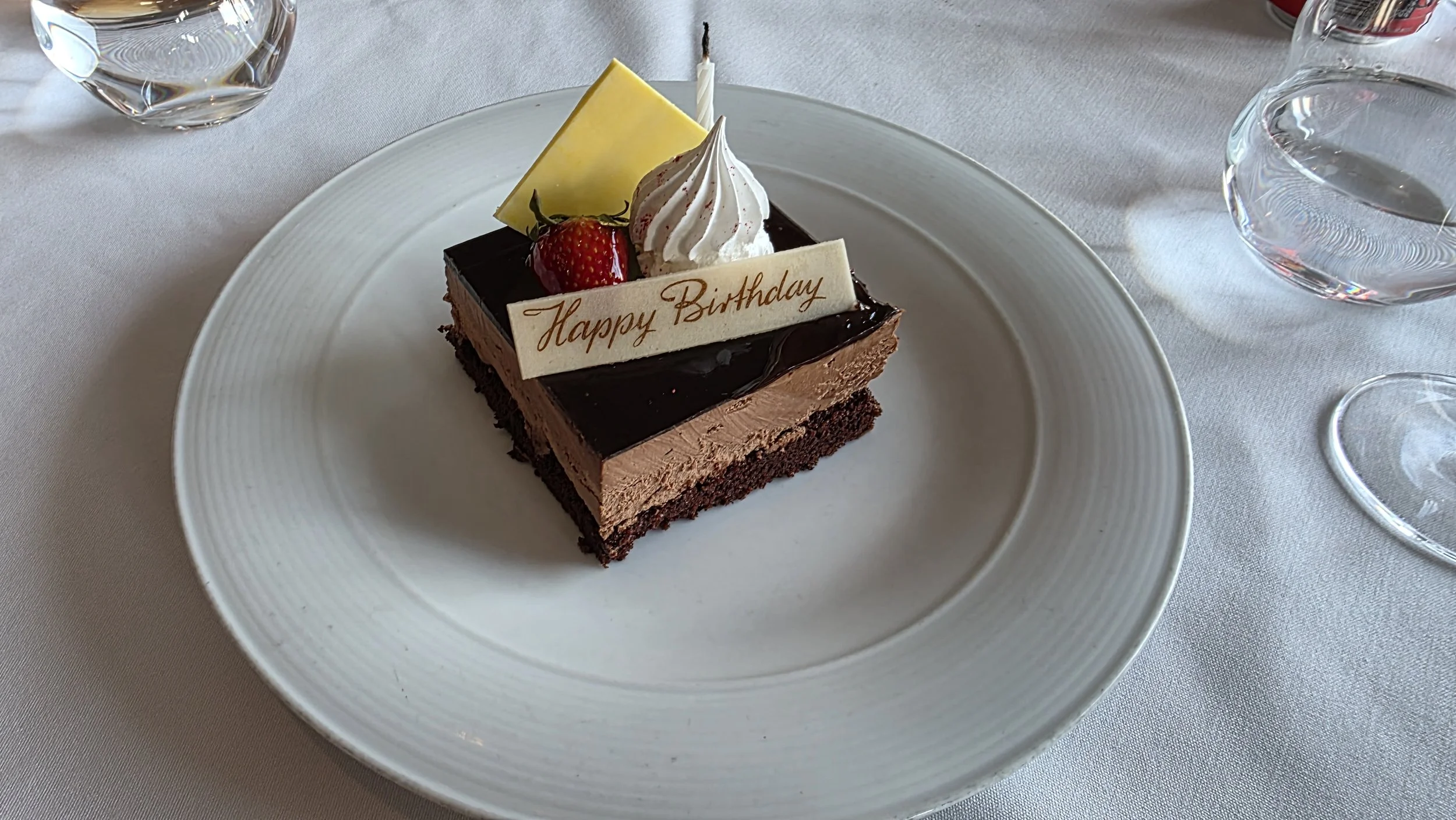 A square chocolate birthday cake with layers of chocolate mousse and cake, topped with a white chocolate piece, a strawberry, whipped cream, and a birthday candle on a white plate.