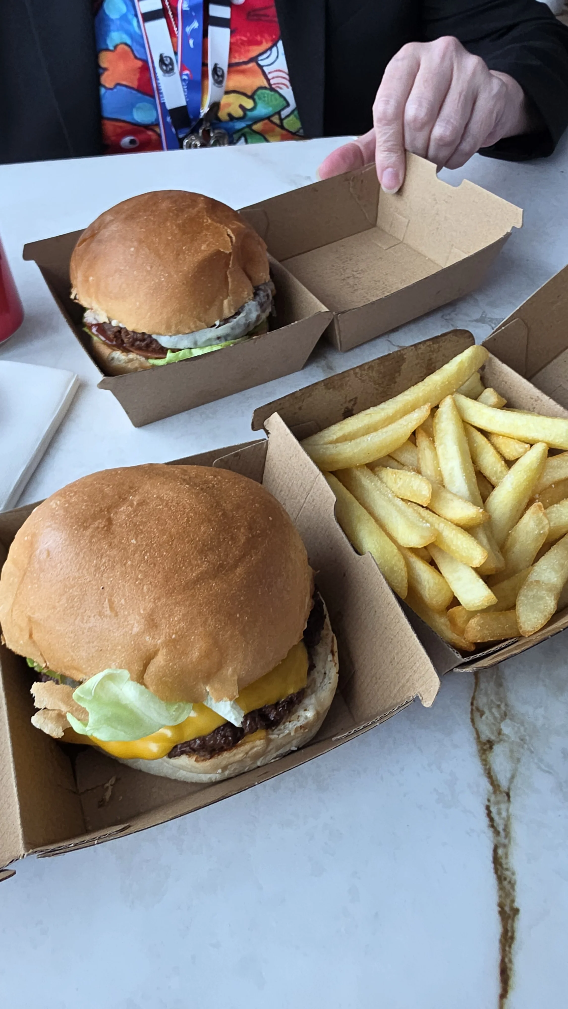 Two cheeseburgers with lettuce, cheese, and beef patties, a tray of French fries, and a person's hand opening the lid of a burger box in a restaurant setting.