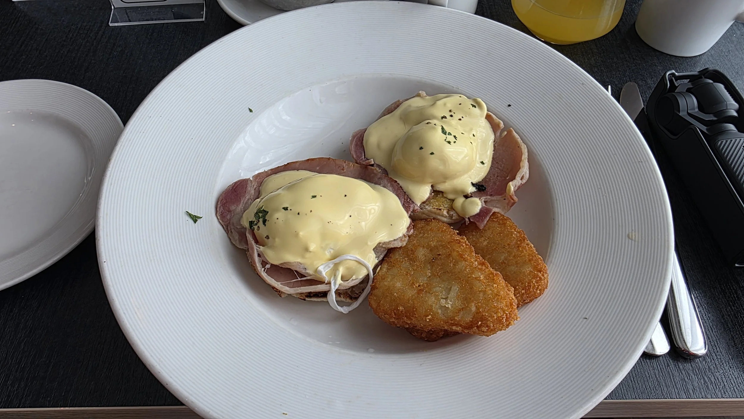 Two eggs Benedict with ham and hollandaise sauce, served with hash browns on a white plate.