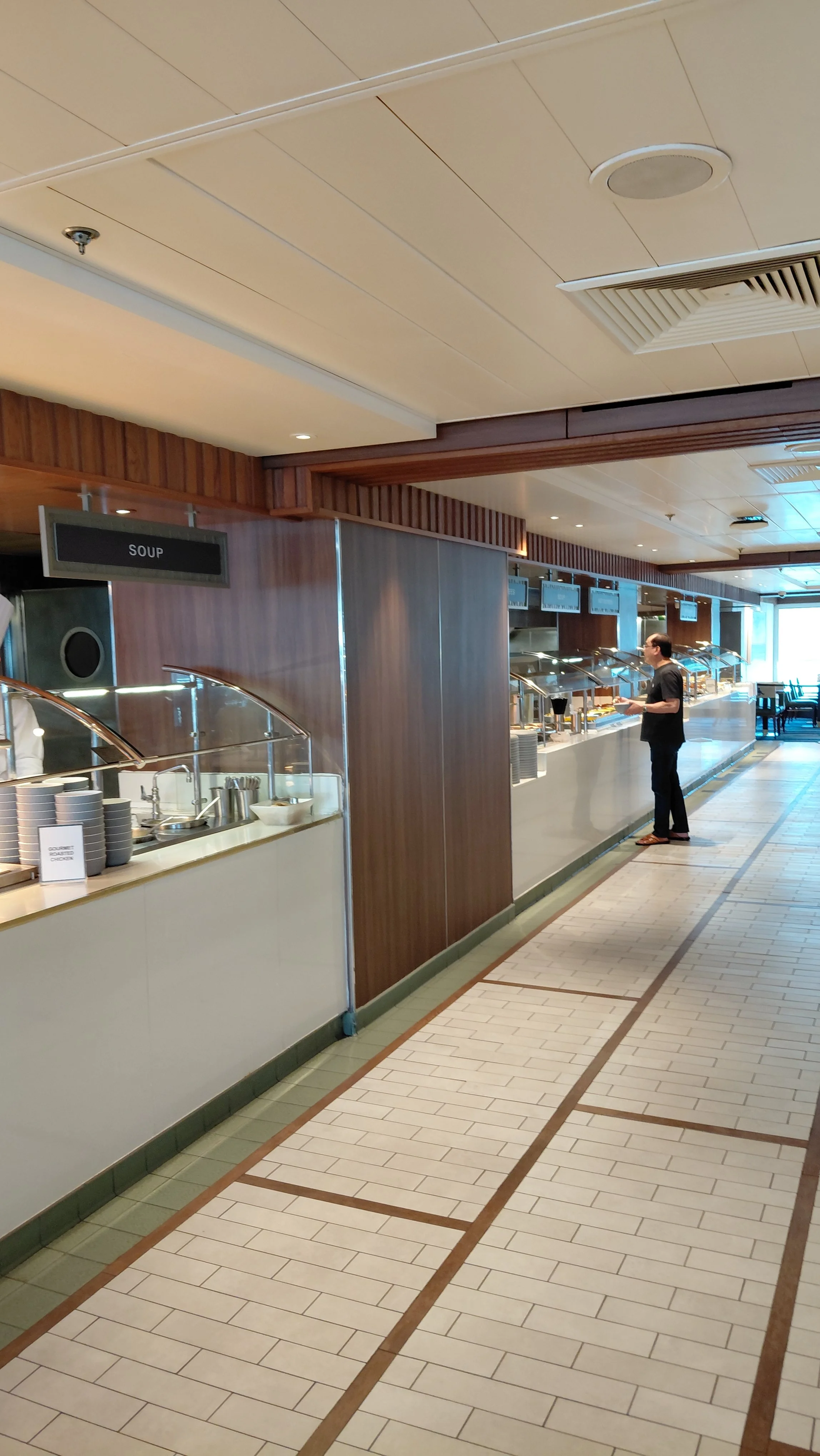 A buffet-style food service area in a restaurant or hotel, with a person serving themselves at the counter. There are various food stations with covered trays and a sign indicating 'SOUP'.