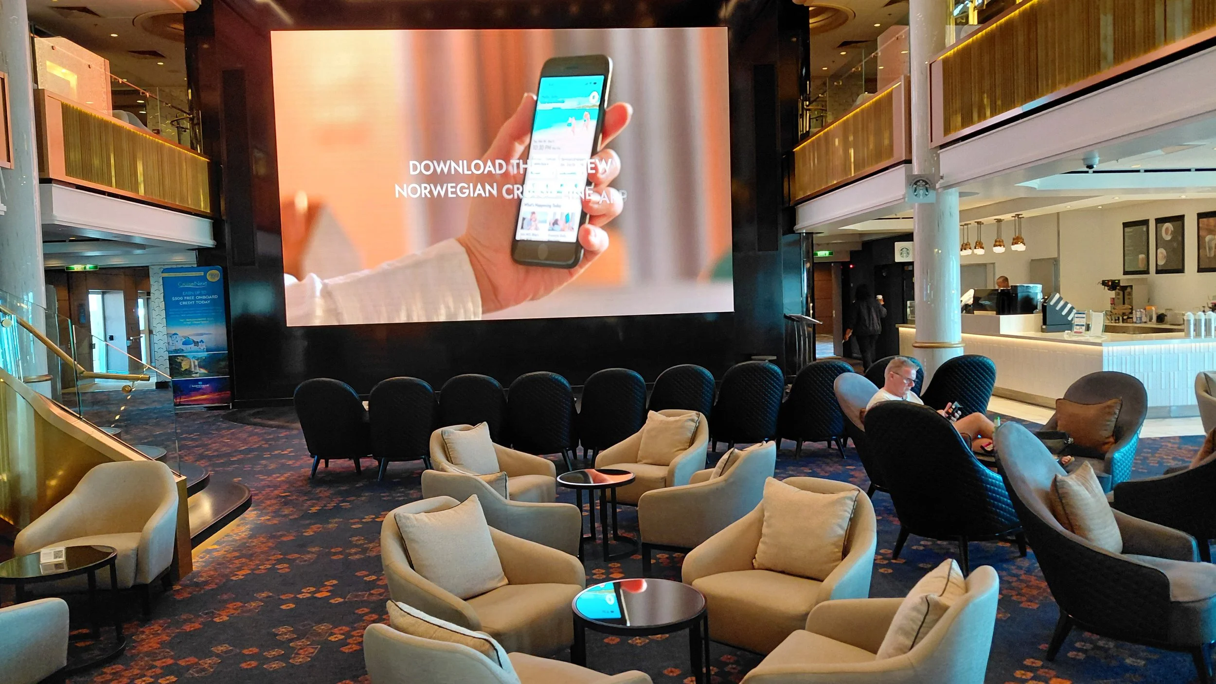 A large screen in a hotel lobby displays an advertisement to download the Norwegian Cruise Line app, with a hand holding a smartphone. The lobby has seating areas with beige and black armchairs, a bar area, and a few people sitting and using their ph