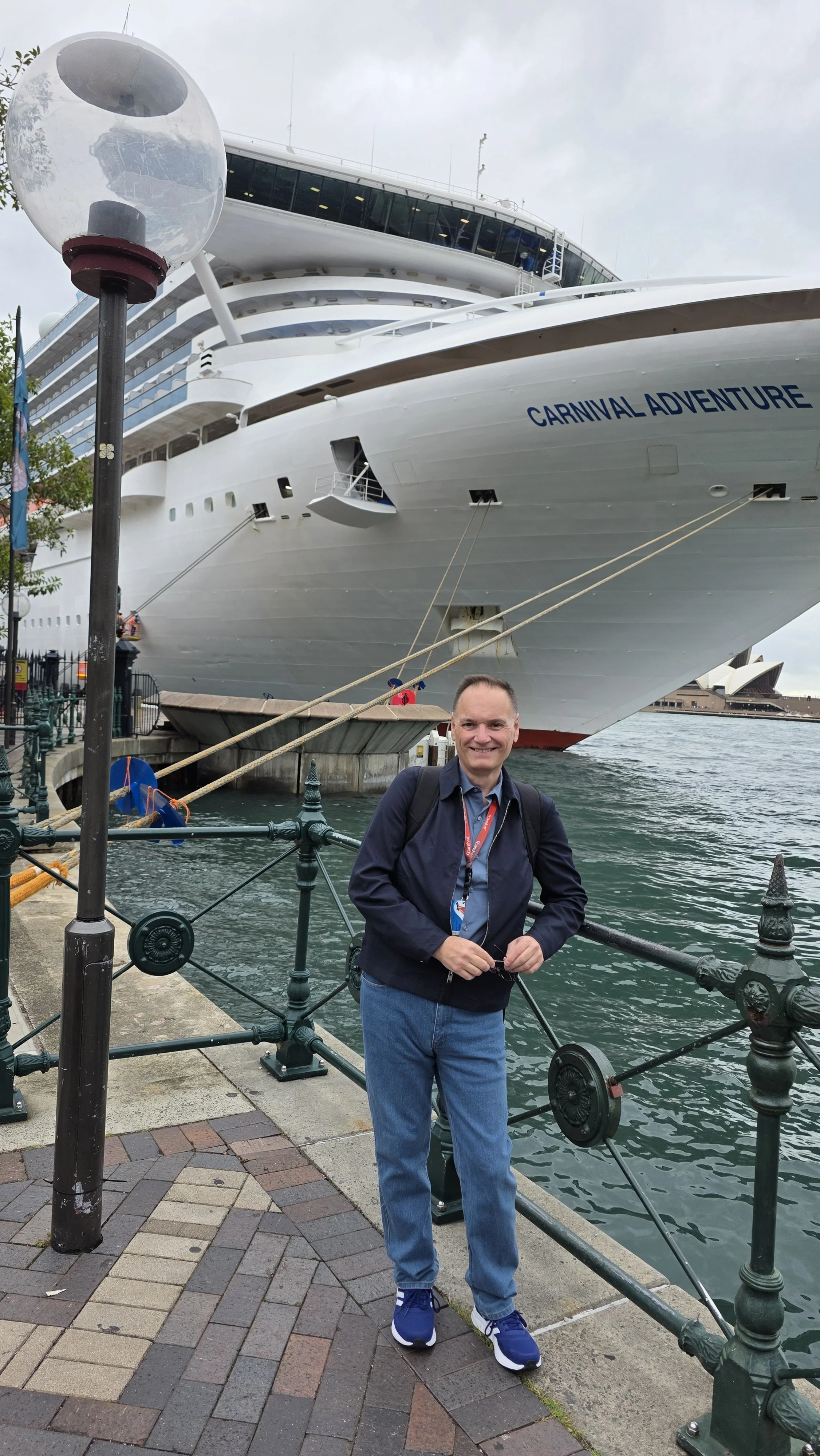 A smiling man standing on a dock in front of a large cruise ship named Carnival Adventure, with water and cityscape in the background.