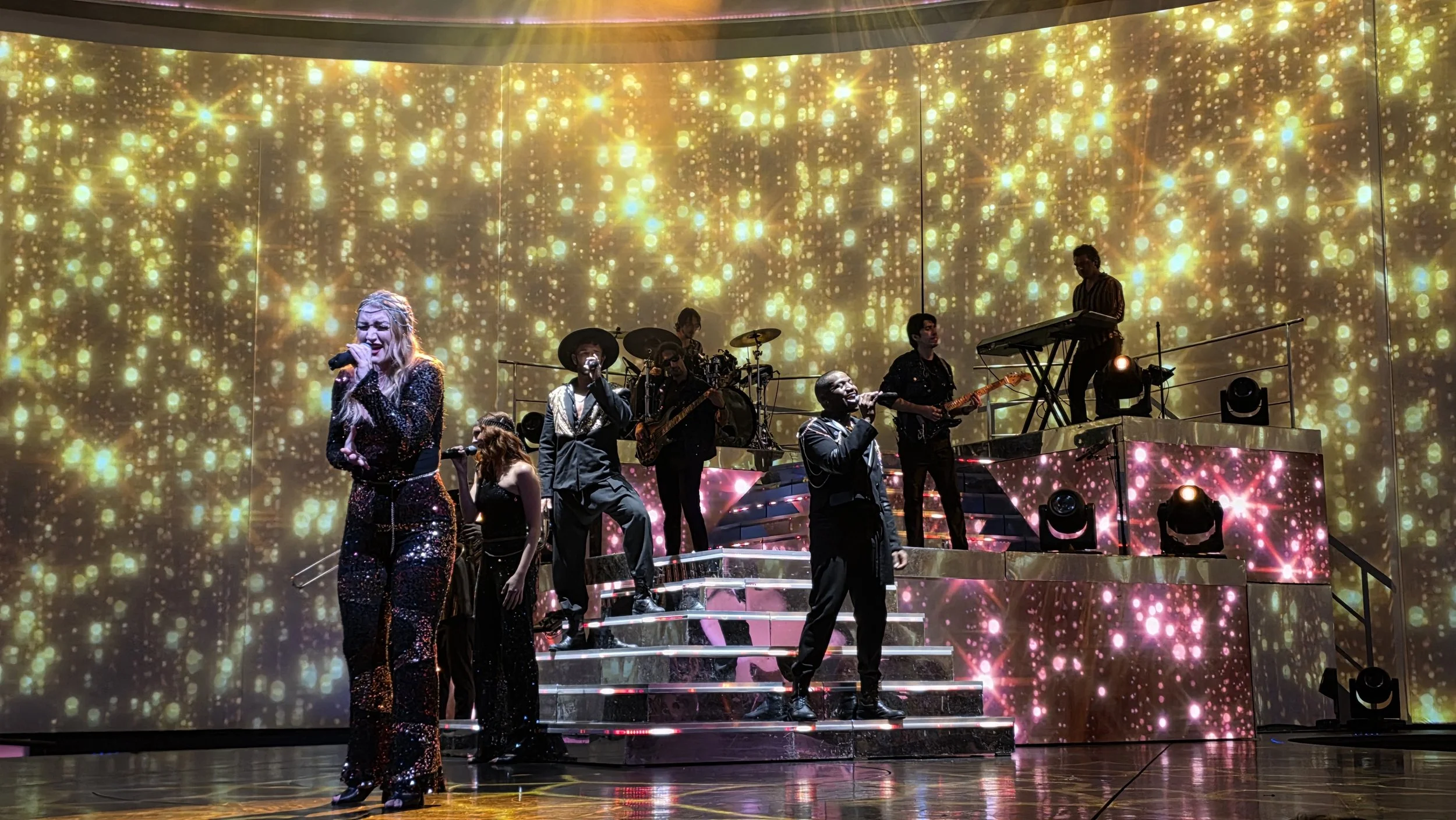 A musical band performs on stage with a backdrop of golden and pink sparkling lights, featuring multiple singers and instrumentalists.