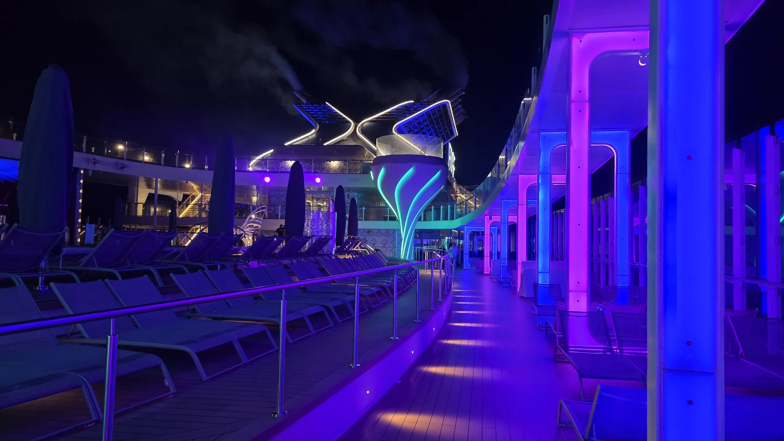 Nighttime scene of a cruise ship deck illuminated with vibrant purple and blue neon lights, with lounge chairs and umbrellas lined along a wooden pathway, and a modern, curved structure glowing in the background.