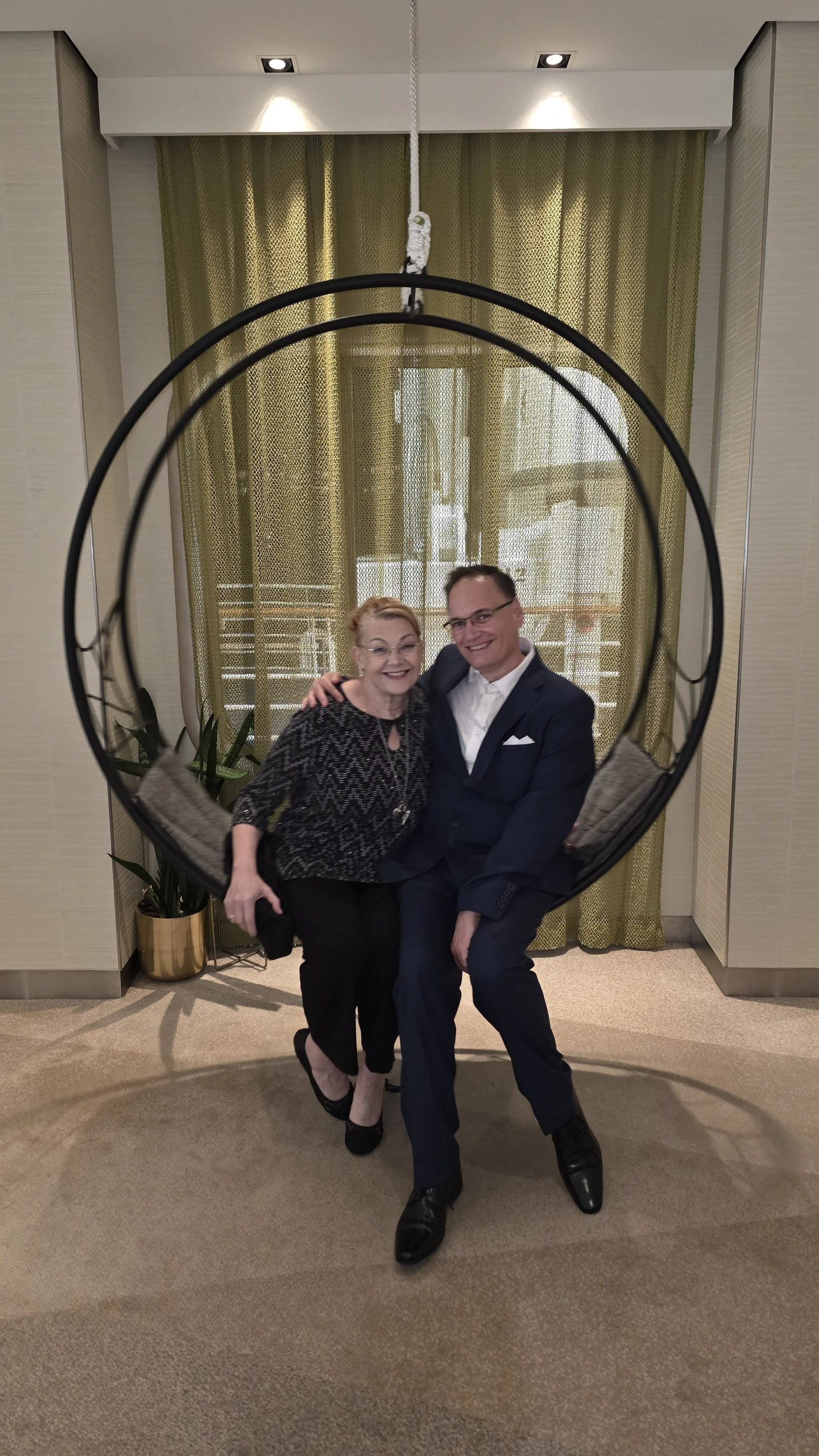 A woman and a man sitting together on a hanging circular swing with pillows, in front of yellow curtains and a large window.