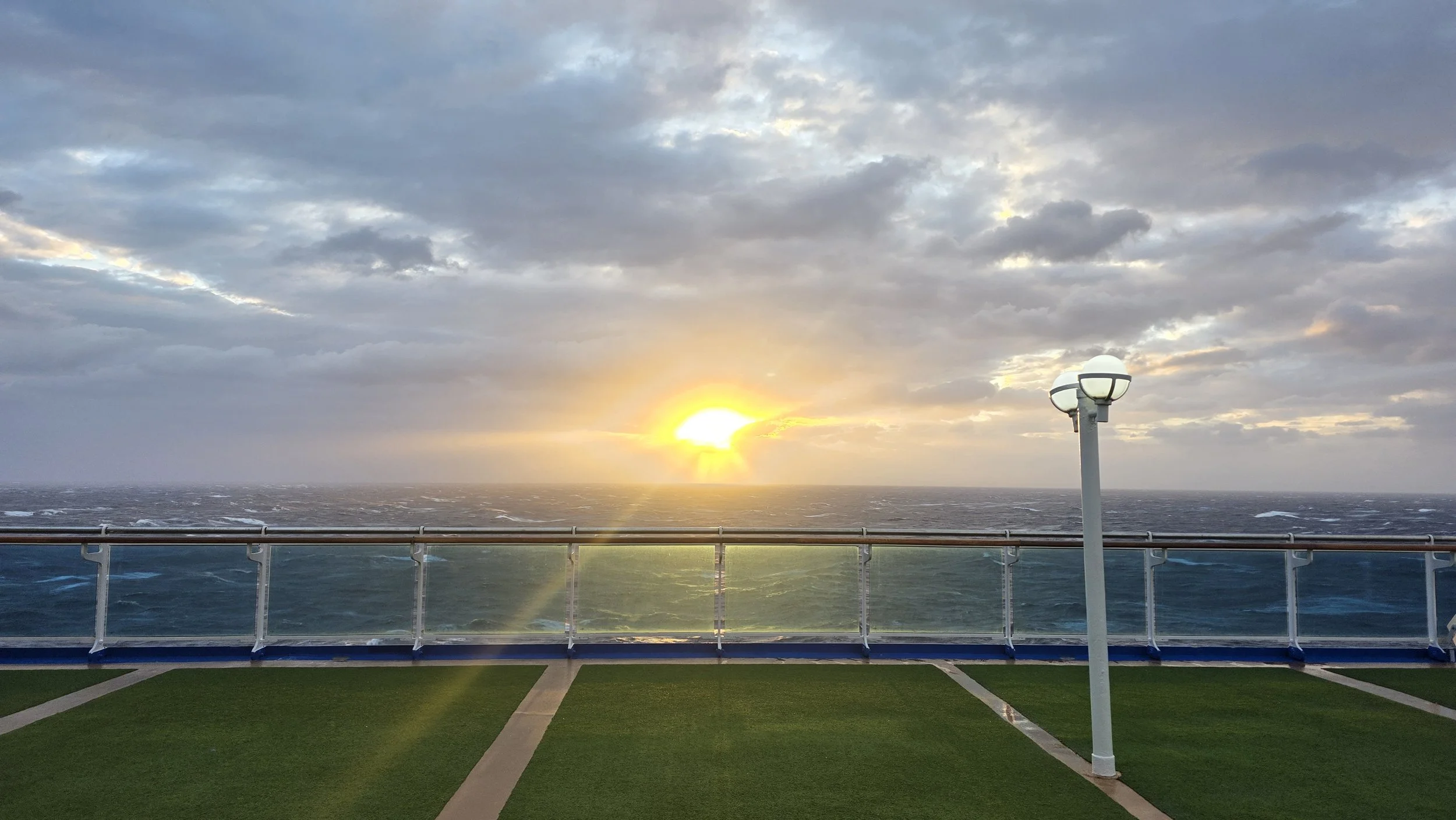Sunset over the ocean viewed from the deck of a cruise ship or coastal building, with artificial grass and a white railing in the foreground.