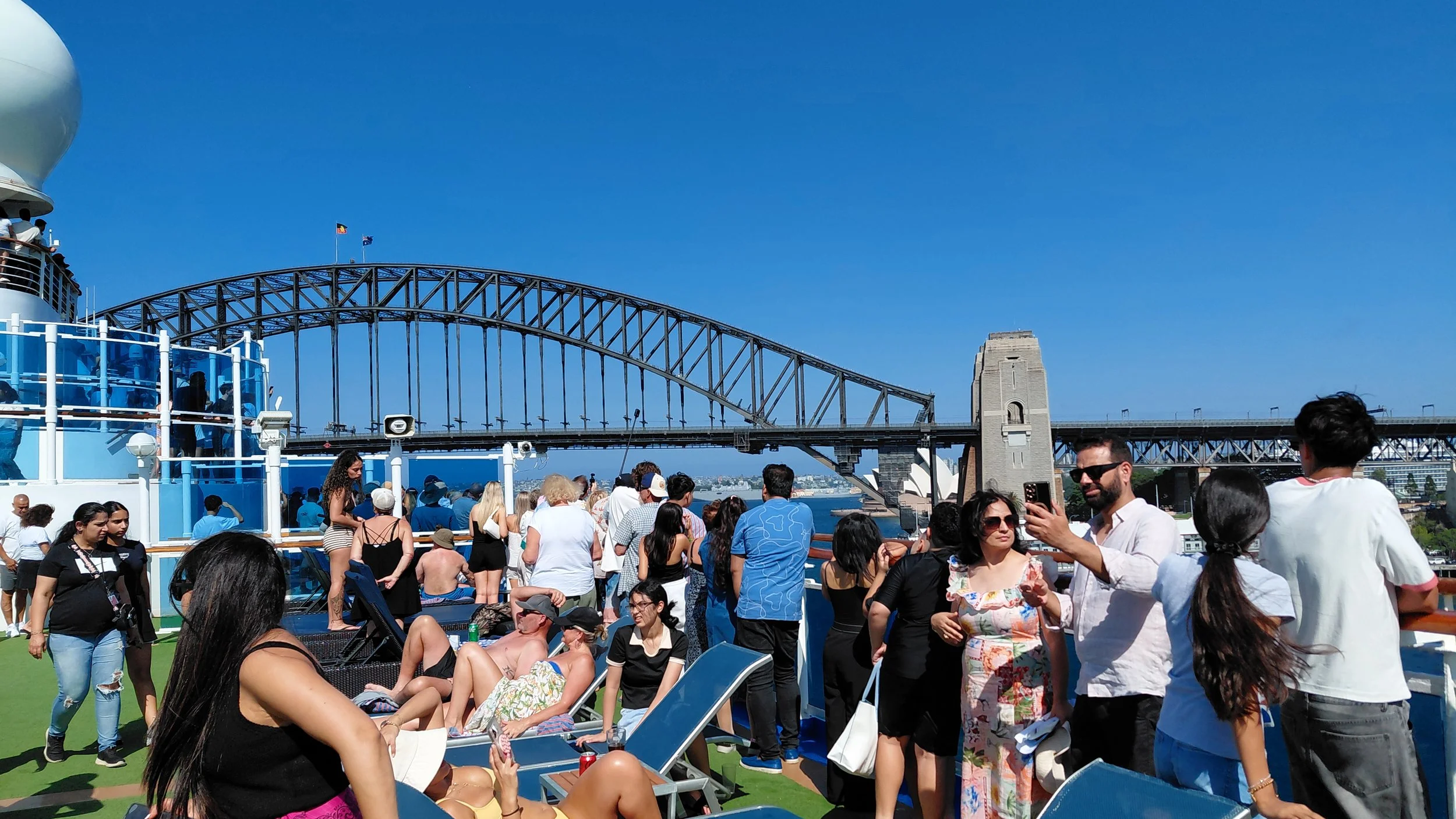 Crowded deck with people relaxing and taking photos, Sydney Harbour Bridge in the background under a blue sky.