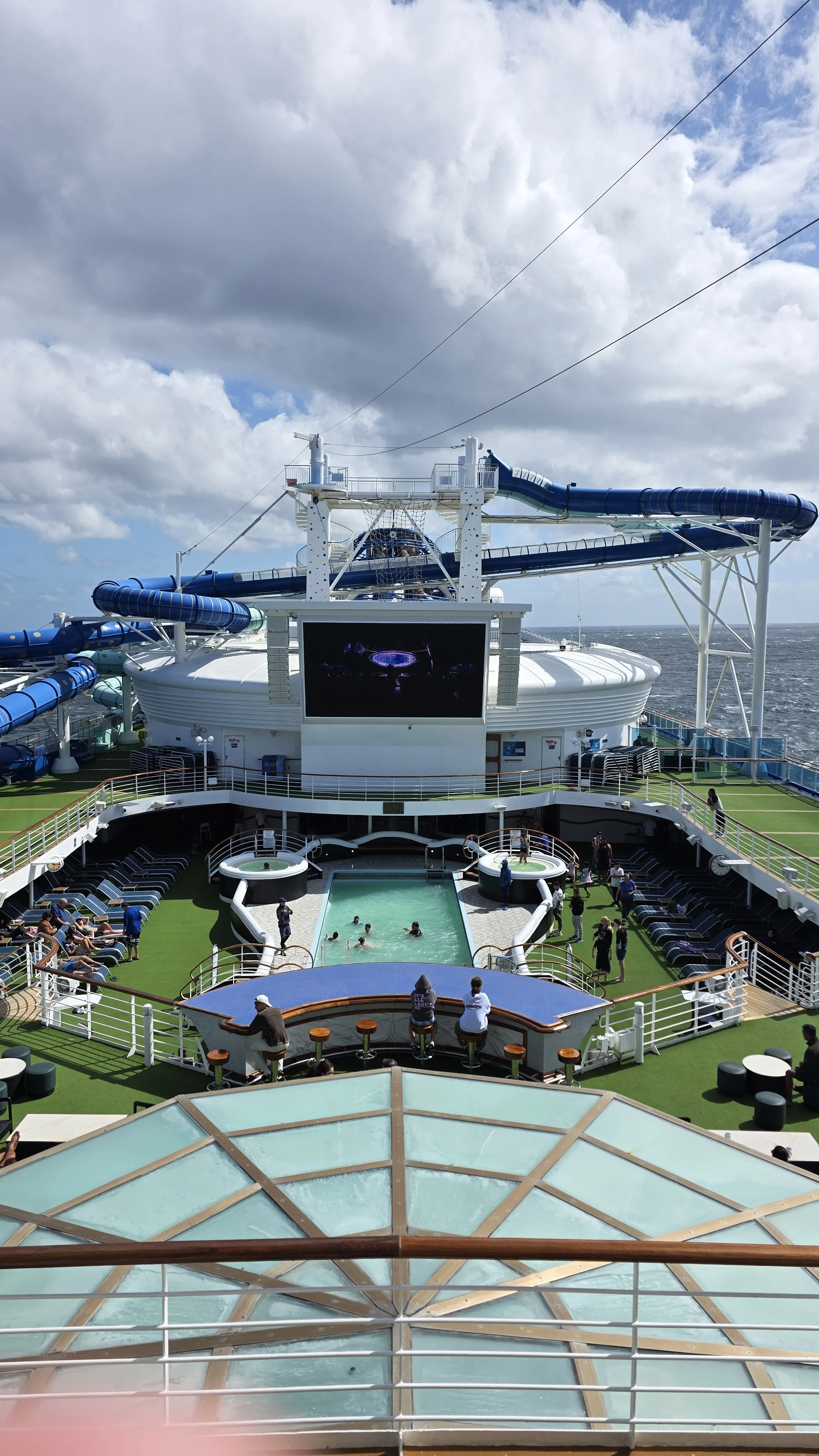 View of the upper deck of a cruise ship with a small pool, lounge chairs, a large screen, water slides, and people enjoying the amenities.