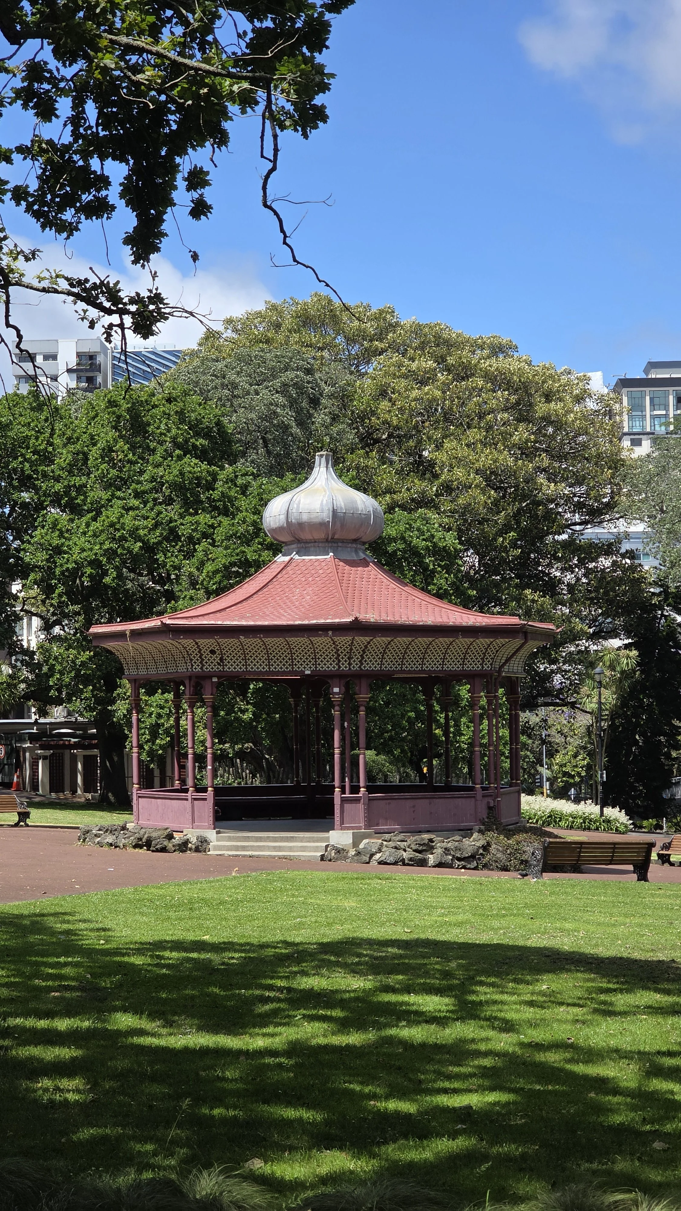 A small, ornate, purple gazebo with a red roof in a park, surrounded by green trees and grass, with blue sky overhead.