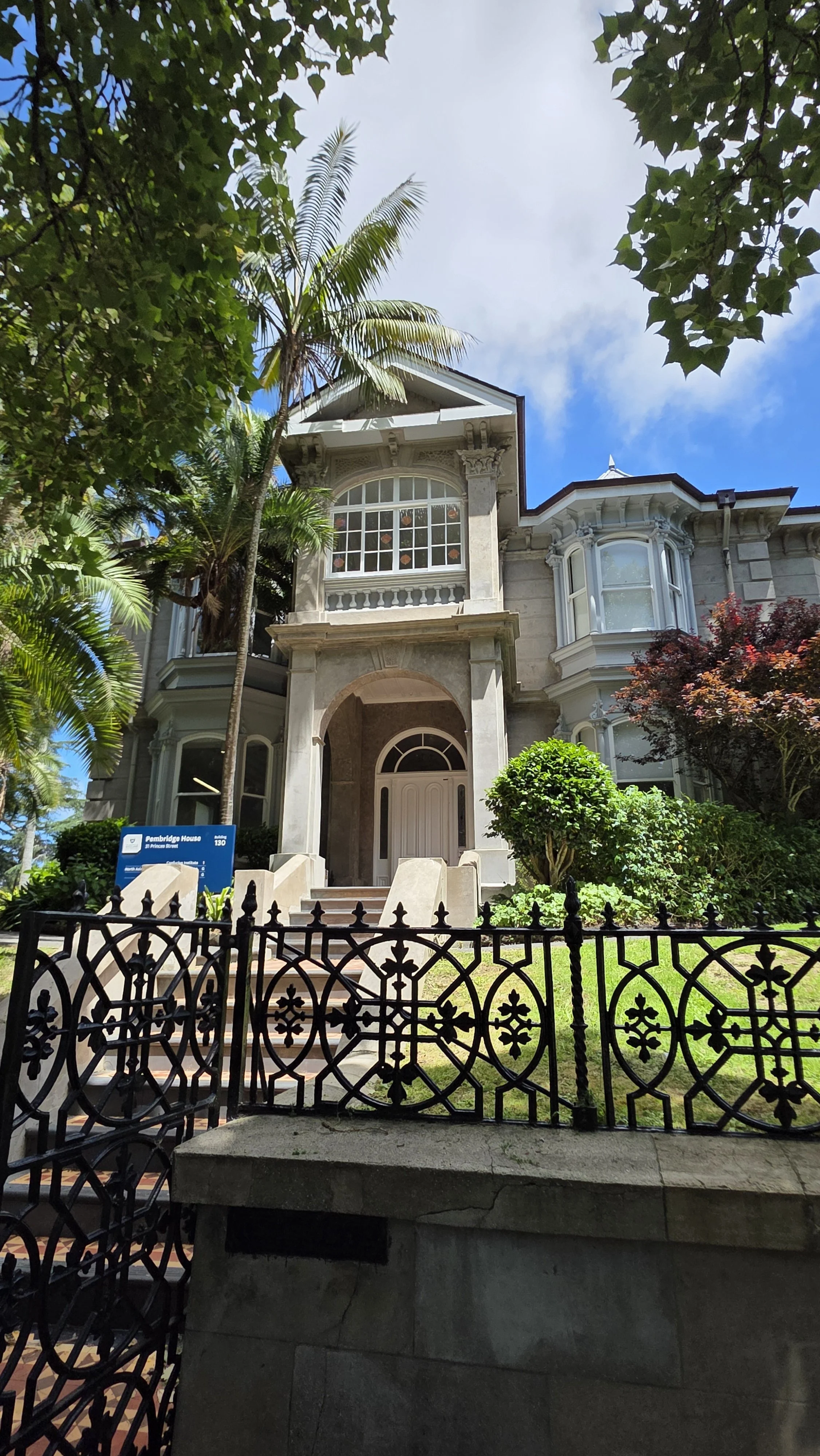 A historic Victorian-style house with a lush garden, featuring a large bay window, front stairs, and an ornate iron fence, with tropical trees and a blue sky in the background.