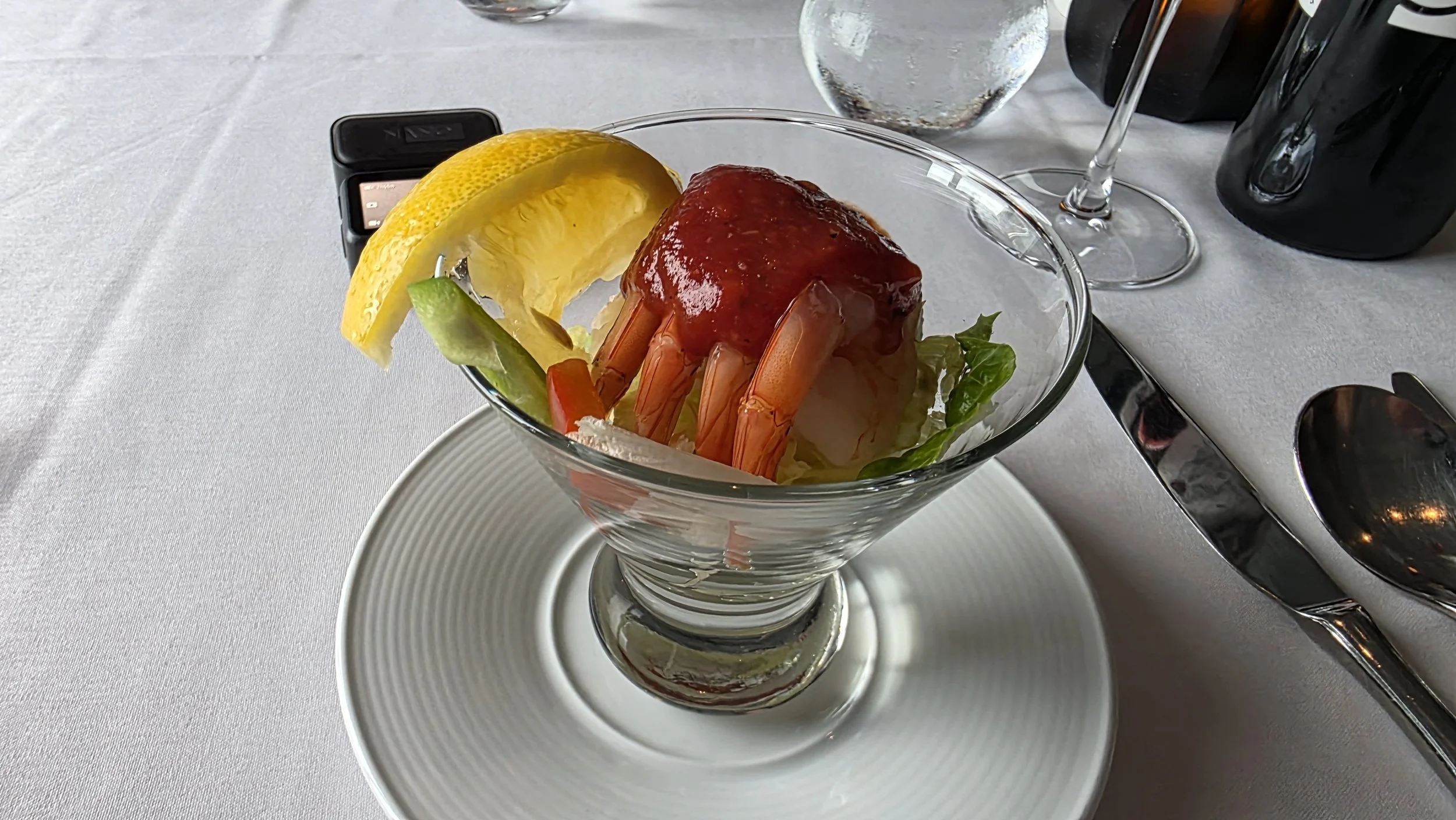 A glass dish containing a cooked shrimp topped with red sauce, garnished with lemon and lime wedges, placed on a white plate with cutlery nearby.