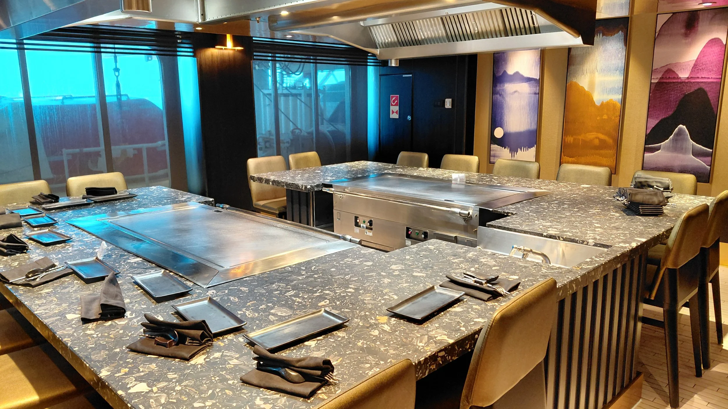 Empty restaurant table setup with black napkins, black plates, and cutlery, featuring a built-in grill in the center, surrounded by beige chairs, with colorful abstract paintings on the wall and large windows in the background.