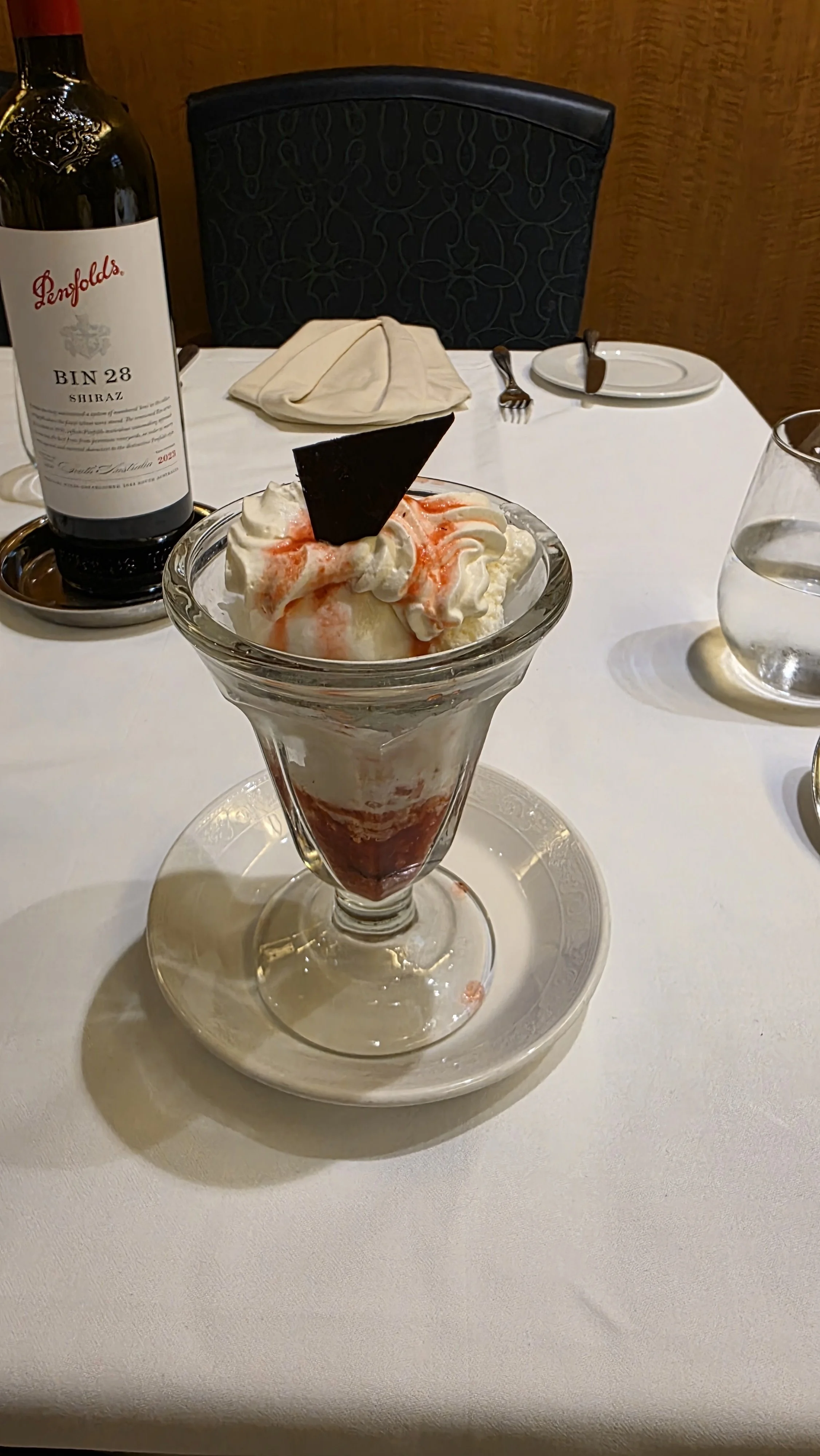 A dessert parfait with whipped cream, strawberry syrup, a piece of chocolate, and strawberry slices in a glass on a white plate on a white tablecloth.