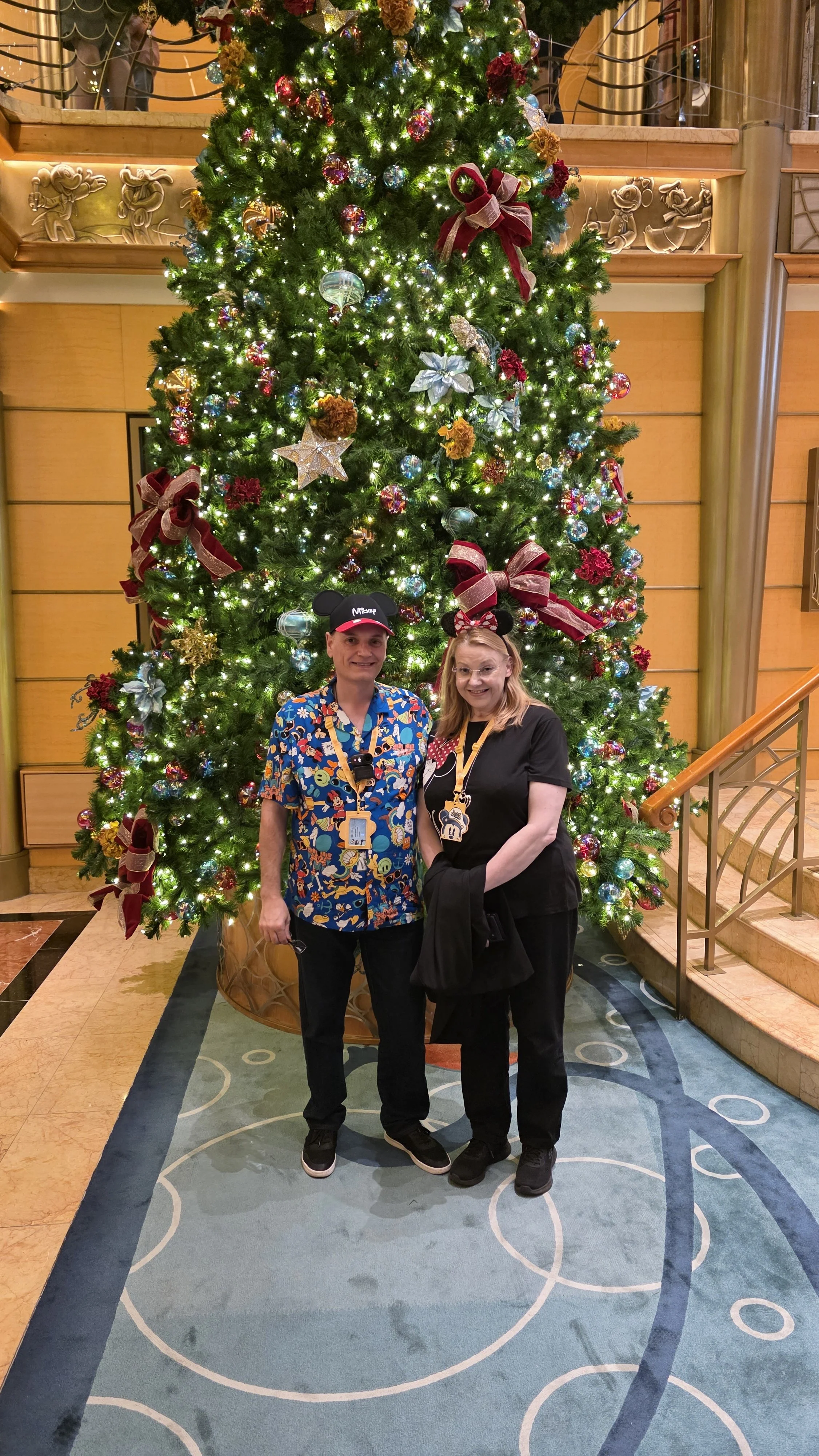 Two people standing in front of a decorated Christmas tree with lights, ribbons, and ornaments in an indoor setting.