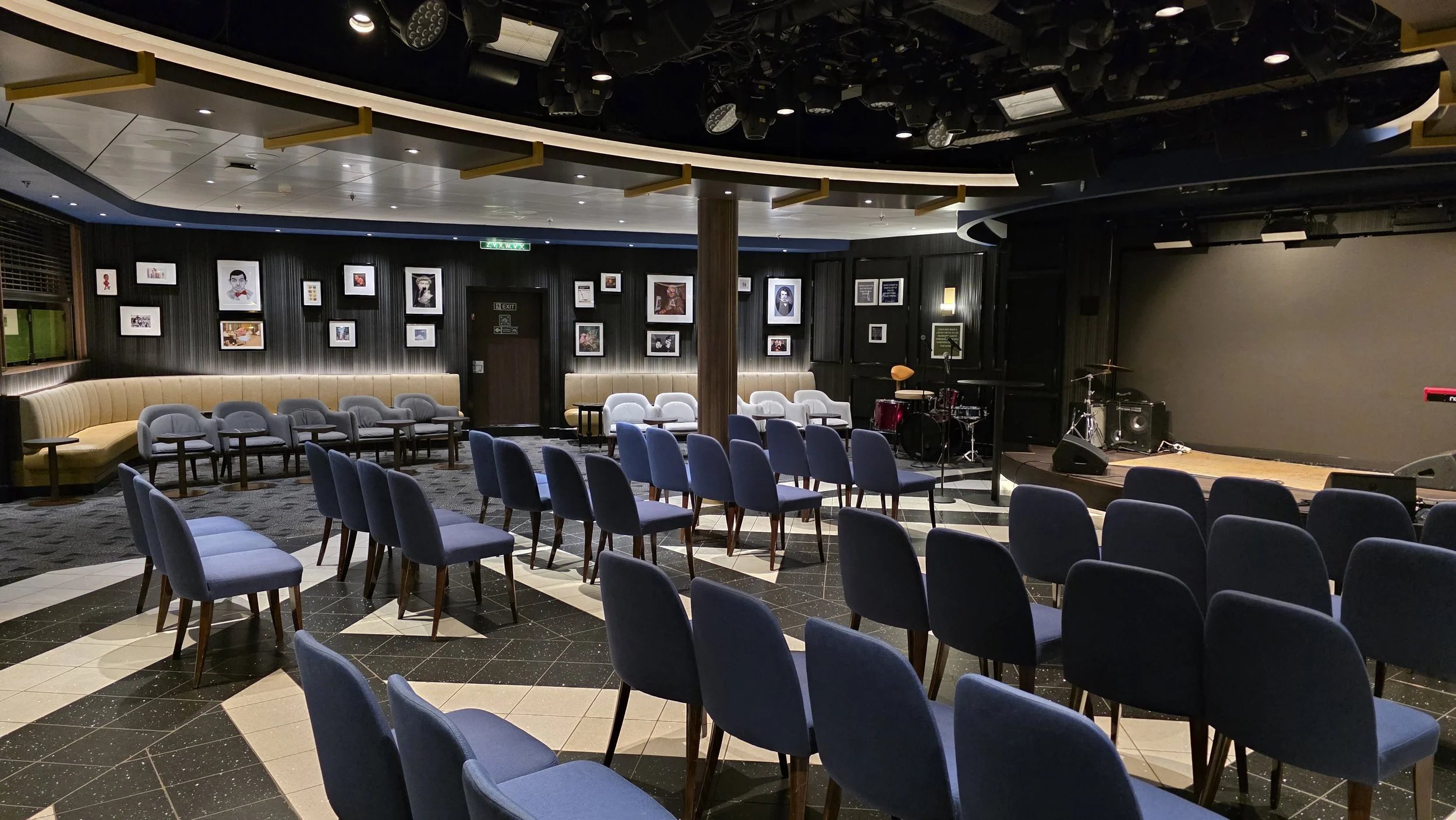 Empty performance venue or small concert hall with rows of blue chairs facing a stage with musical instruments, framed pictures on dark walls, and a neon exit sign.