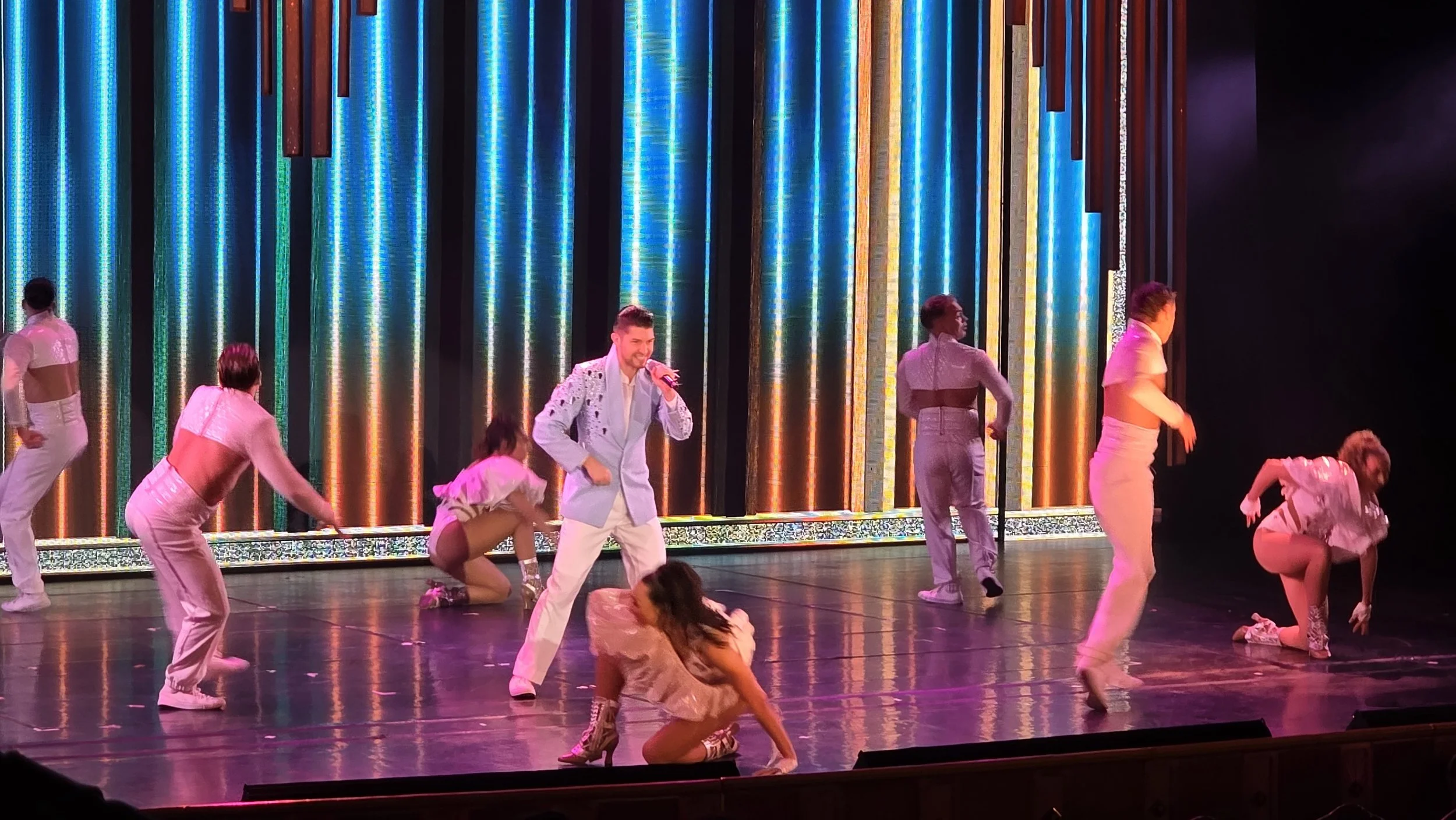 Performers on stage during a dance show with colorful vertical light bars in the background, some dancers crouching or standing, and a male singer holding a microphone in the center.
