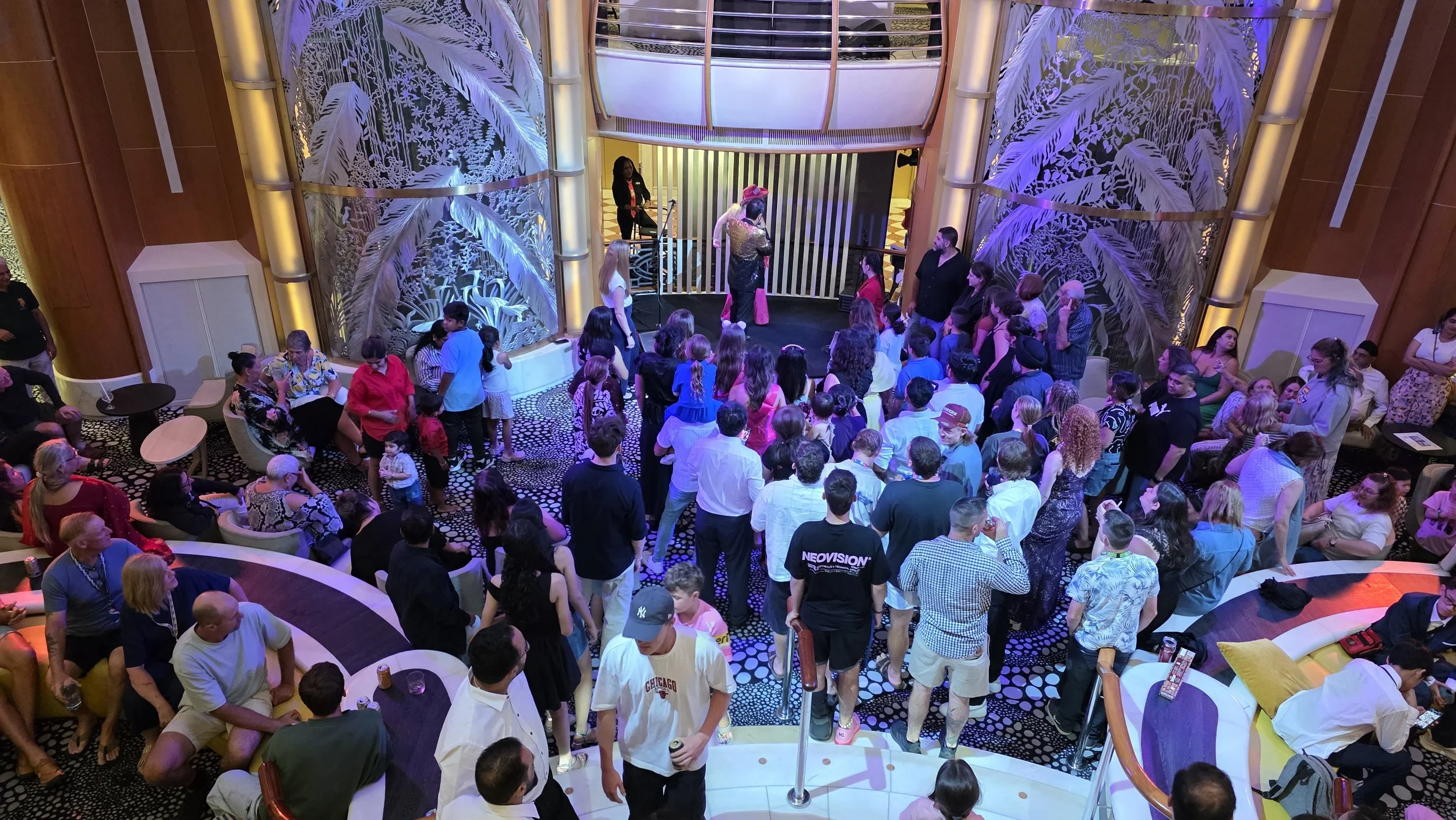 People gathered on a stage and dancing area in a large, decorated theater with jungle-themed wall art, many children and adults watching and participating in a social event or party.