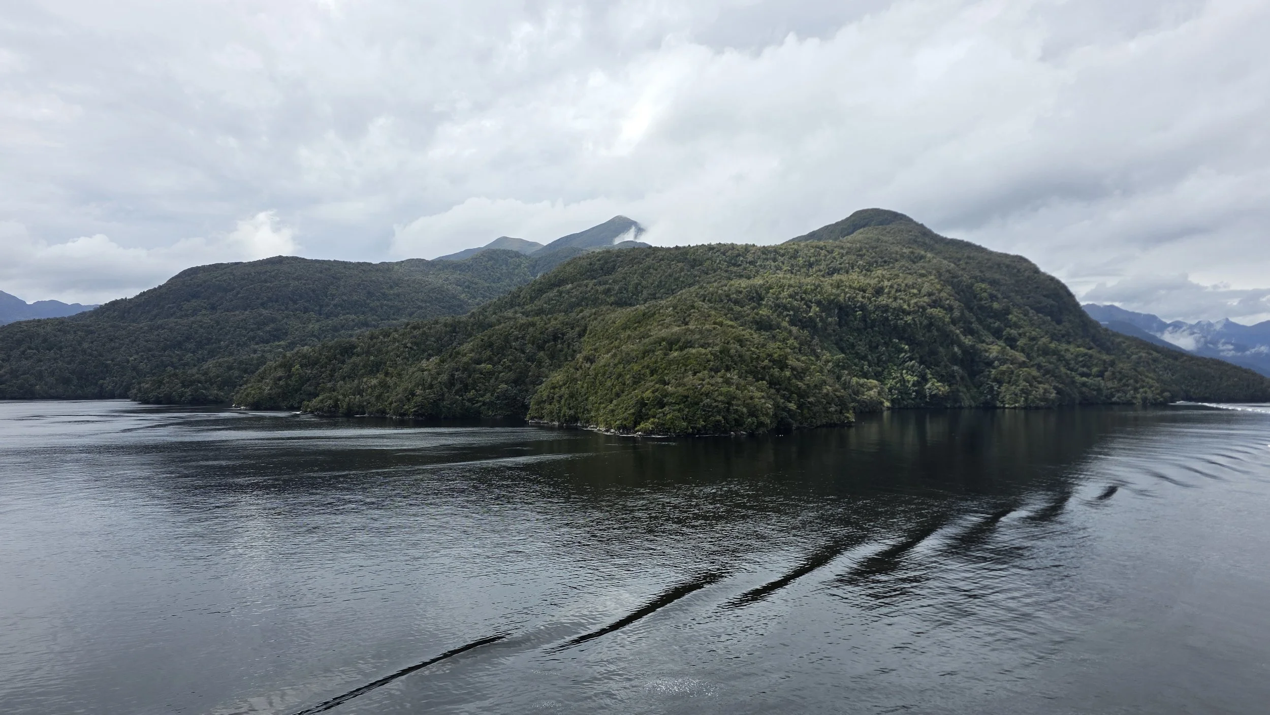 A large, forested island in a body of water with mountains and cloudy sky in the background.