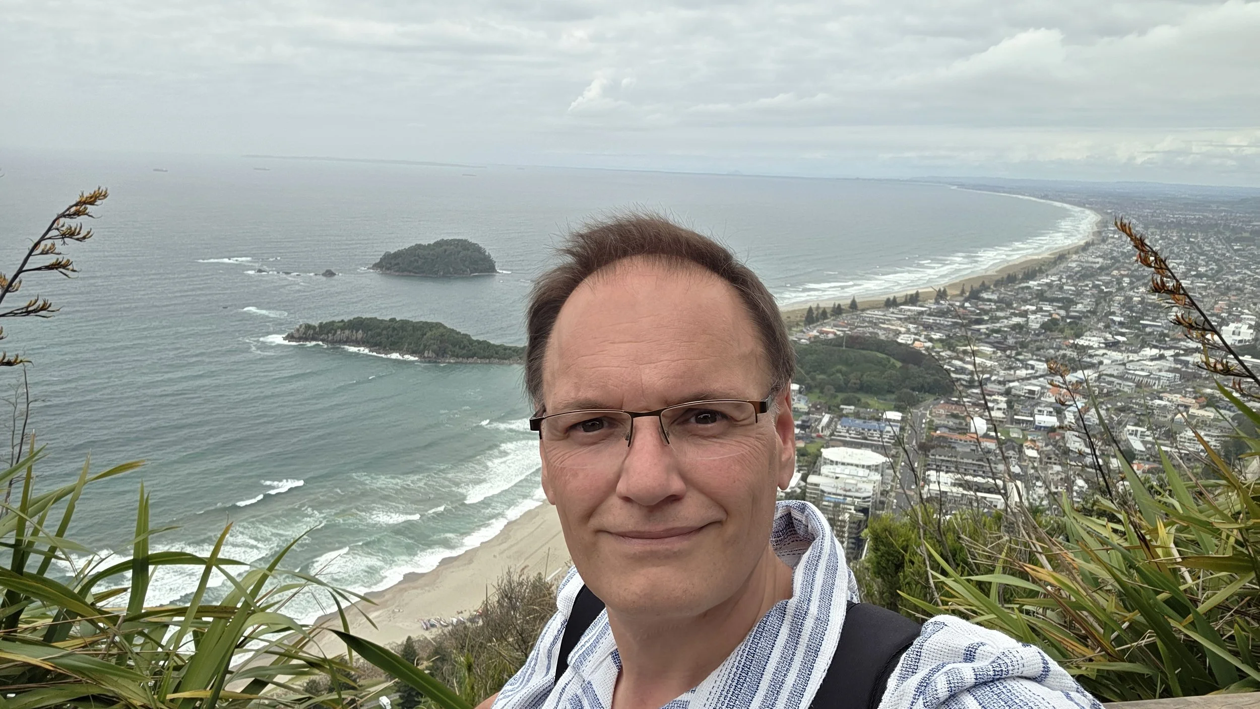 A man taking a selfie on a hillside overlooking a beach, ocean, and a city at the coast with two small islands nearby, under an overcast sky.